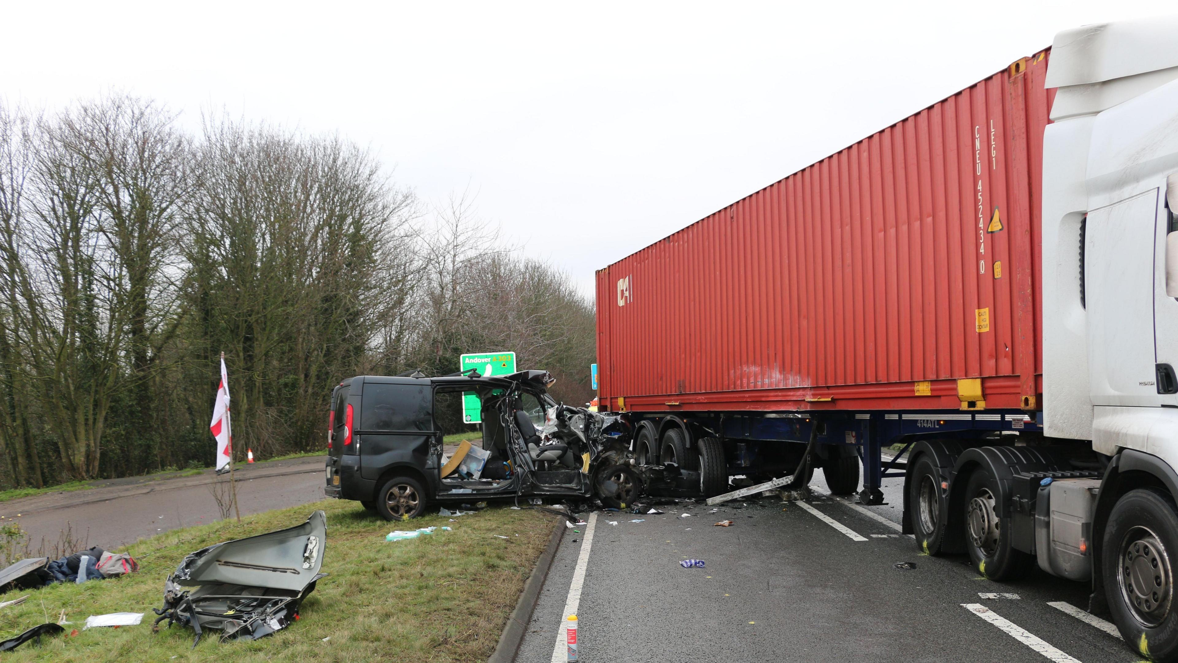 A black van is pictured crashed into the rear of a large red lorry. The van is on a verge facing into the road and its front is completely destroyed against the end of the of the sid of the lorry. Debris is littered nearby. 