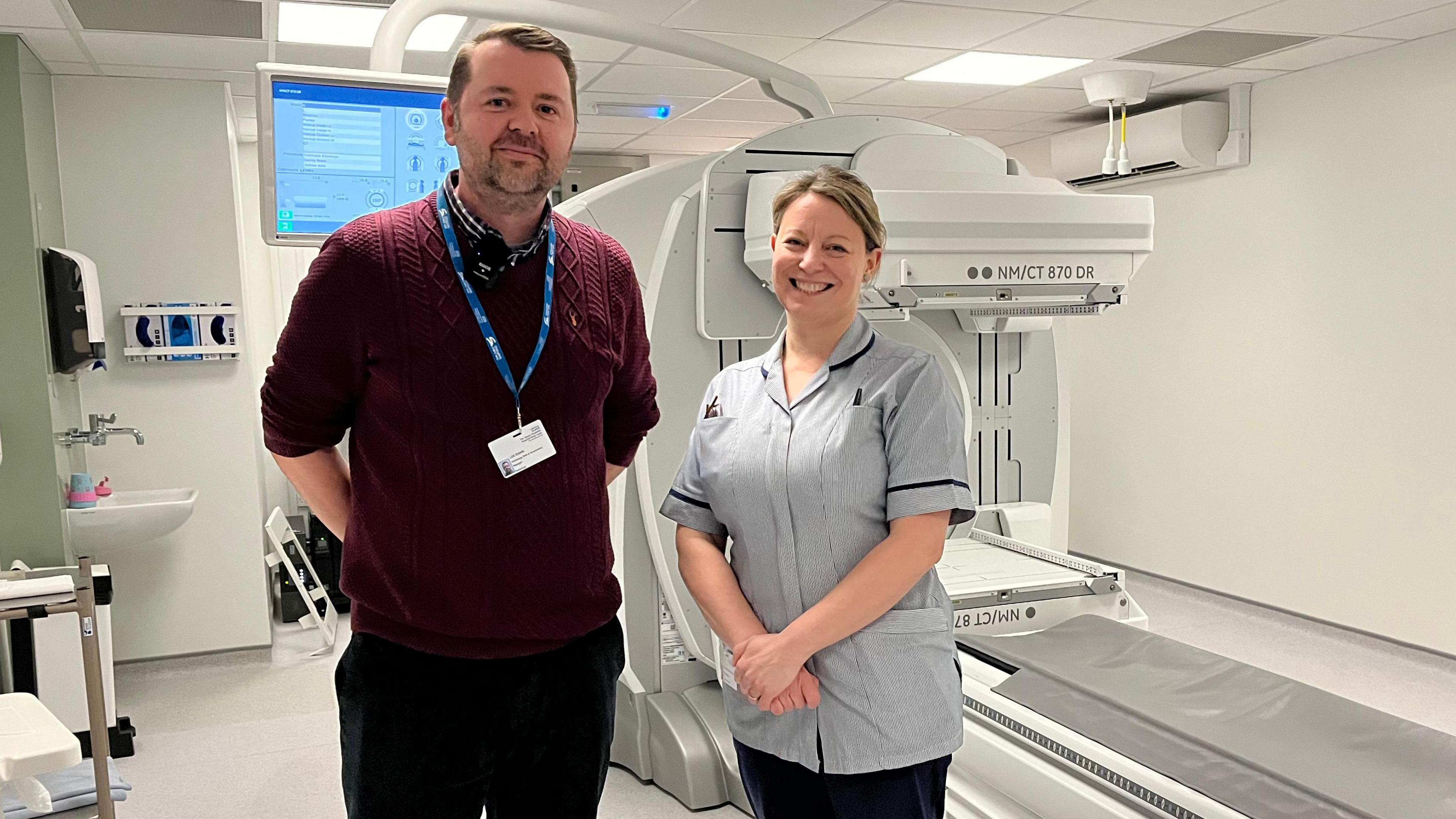 Lee Evans is Radiology Risk Manager at the Queen Elizabeth Hospital pictured with Hannah St Barbe Oldham, Nuclear Medicine Lead Radiographer. Lee is wearing a crimson knit jumper and black trousers. Hannah wears light grey scrubs and black trousers. 