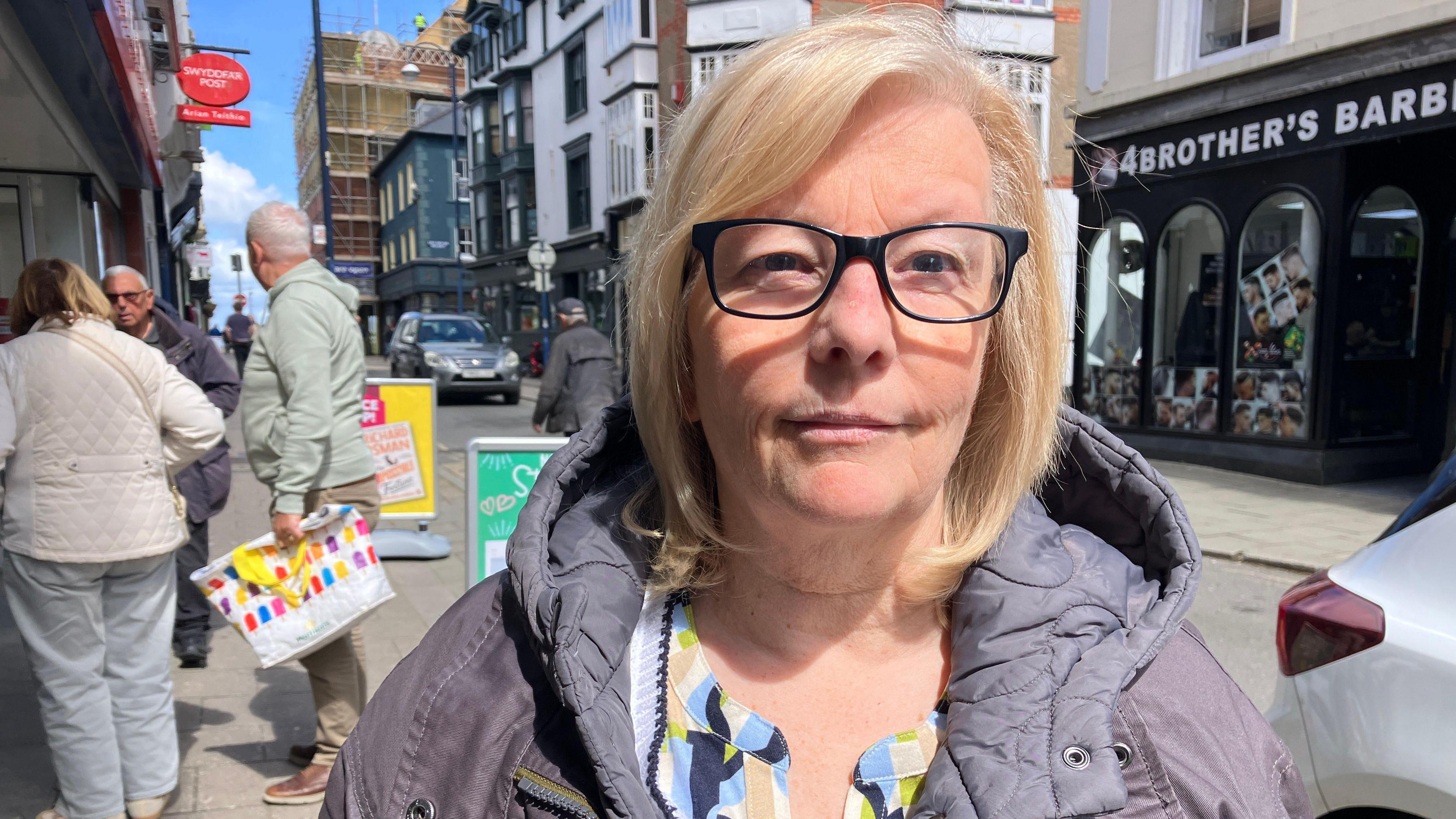 Llun o Lynn Phillips yn sefyll yng nghanol y dref yn Aberystwyth - sbectol a chot lliw piws