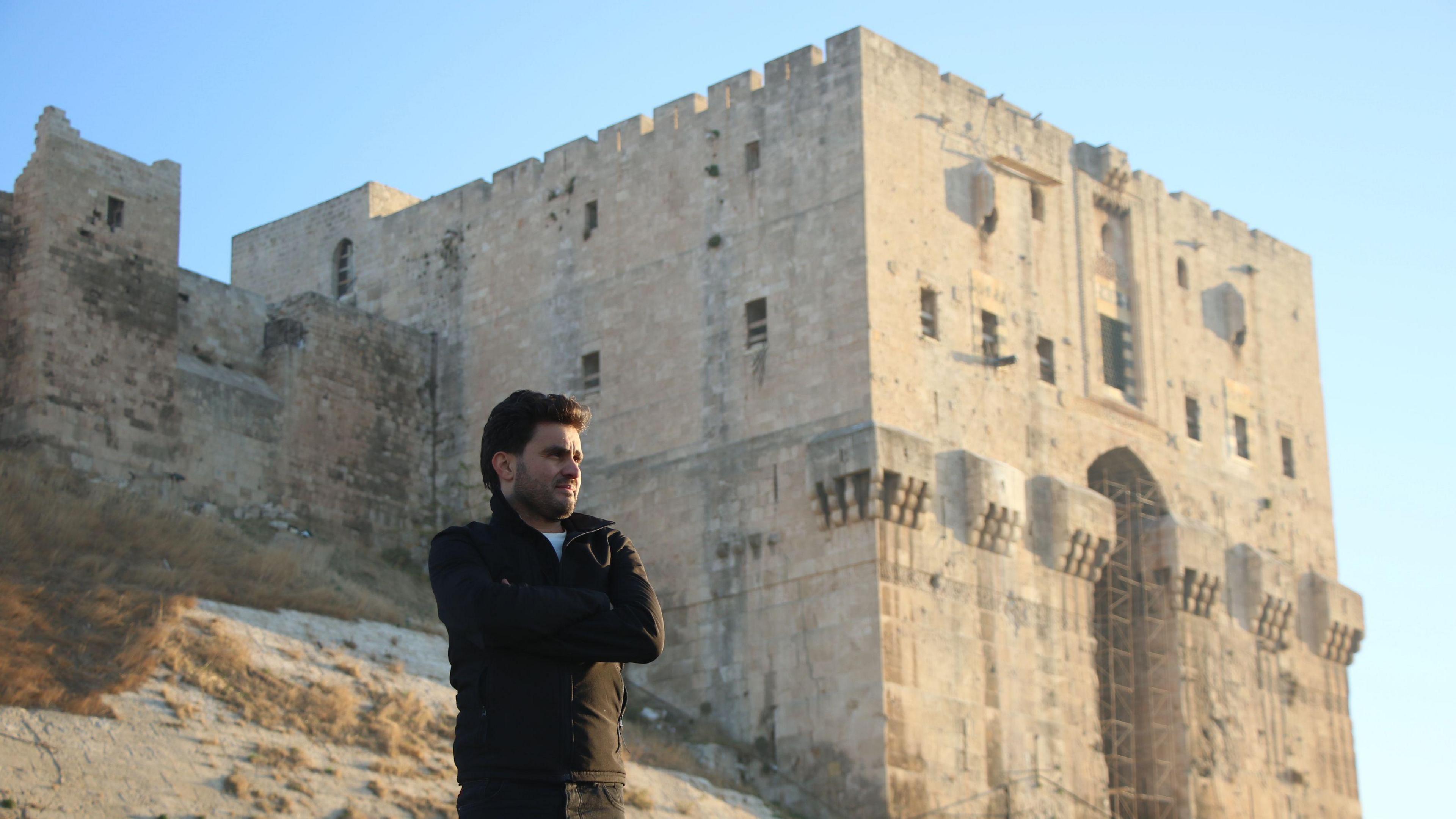 Abdulkafi stands in front of the Aleppo Citadel, in the heart of the Syrian city. He is wearing black and folding his arms. 