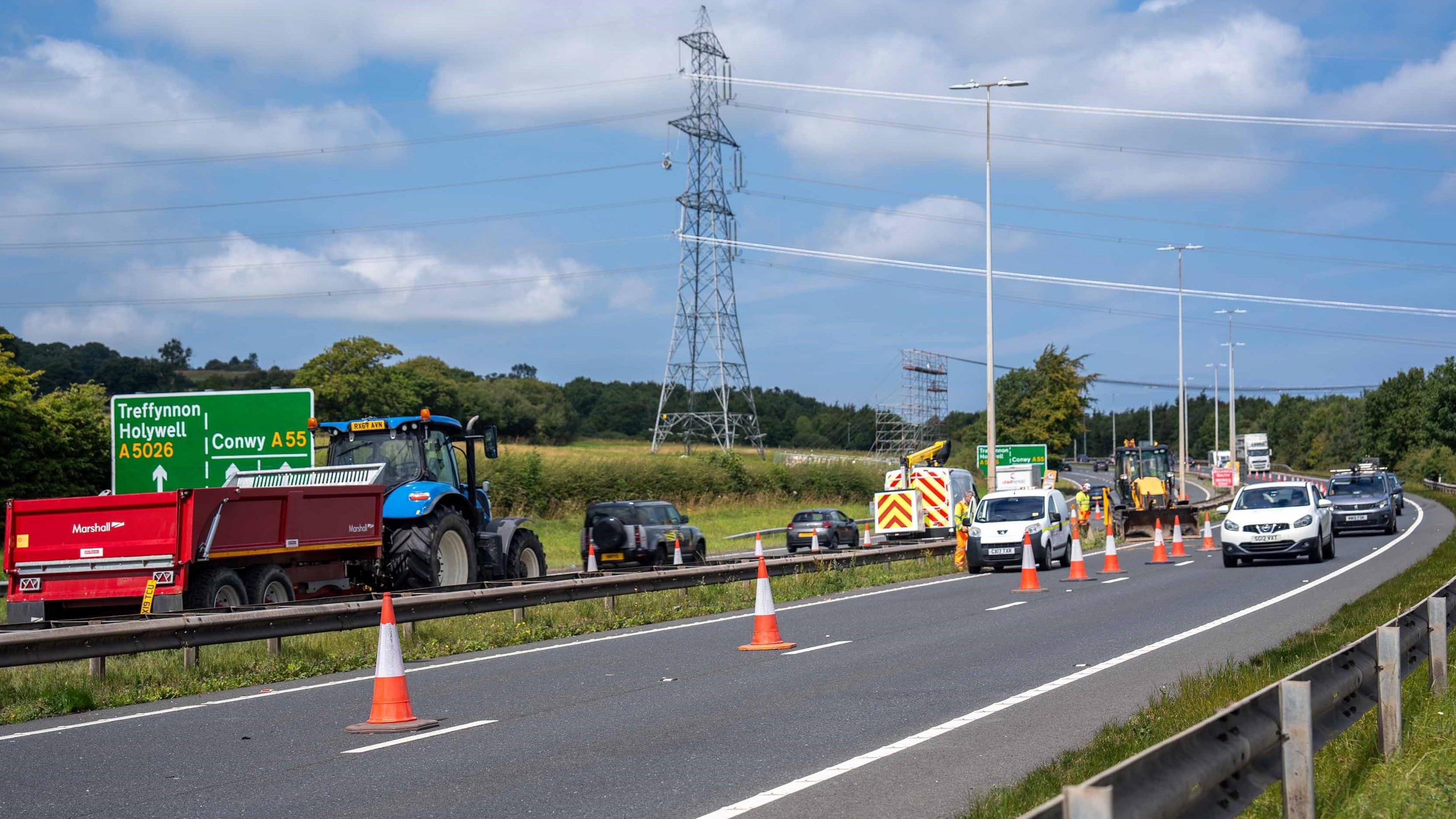 A busy dual carriageway with lots of vehicles, with only a single lane open on each side and the other filled with orange cones. In the middle are a number of vehicles such as a digger and a works van, and in the background are road signs, fields and an electricity pylon.