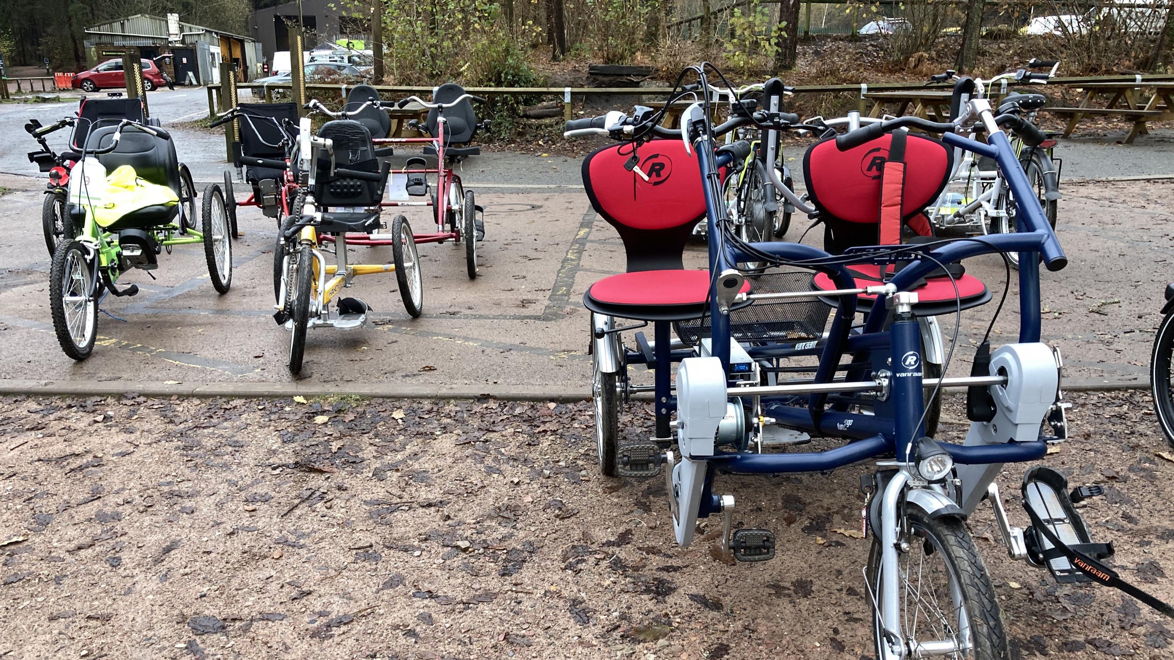 Seven of the adapted bikes lined up on the muddy ground in the Forest of Dean. Some of them are side-by-side bikes, some have platforms on the back for securing a wheelchair and some are pedalled by hand.