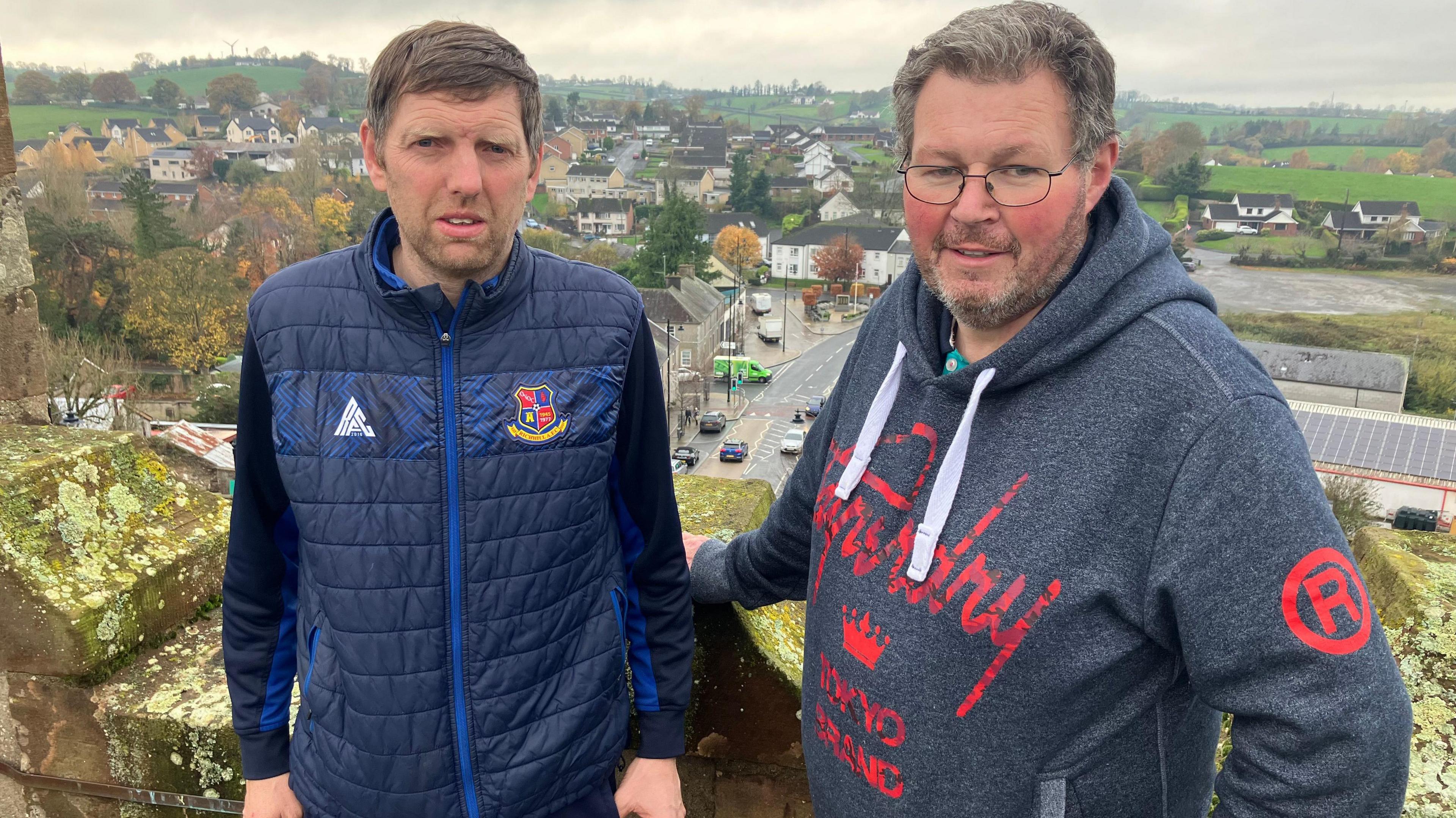 Two men pose with fields behind them. They're standing on a tower.