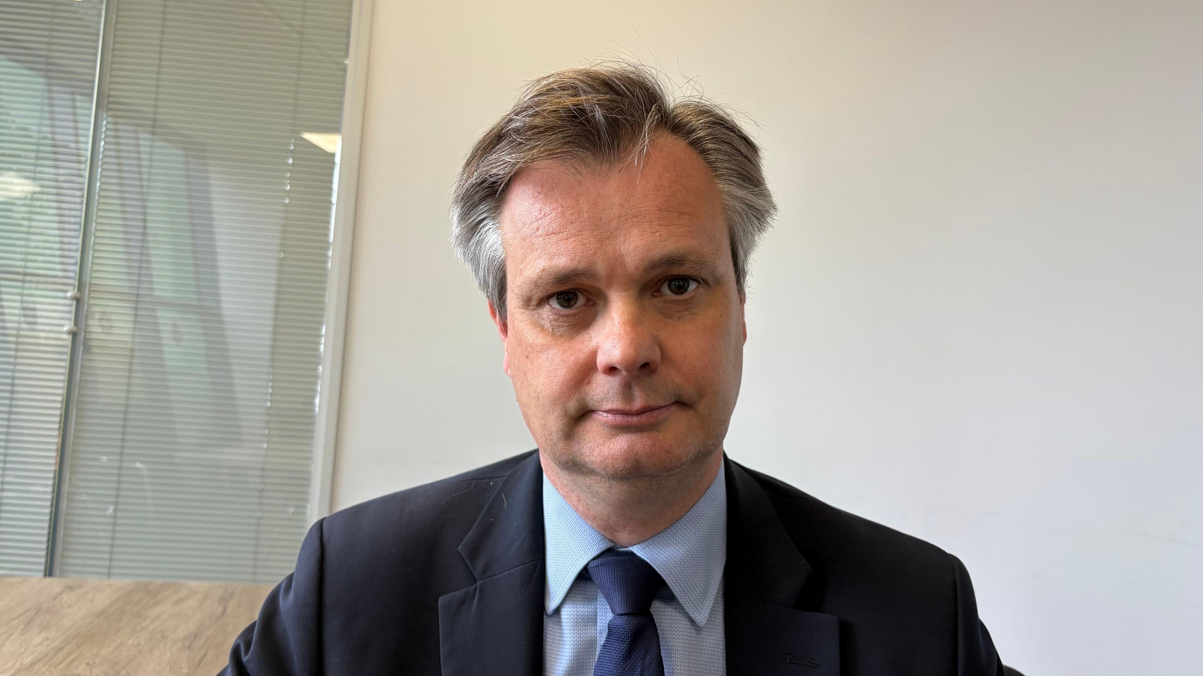 Simon Rouse with medium-length dark hair looking at the camera and wearing a dark grey jacket, light blue shirt and dark blue tie. He is sitting in front of a white wall with a window to the left.