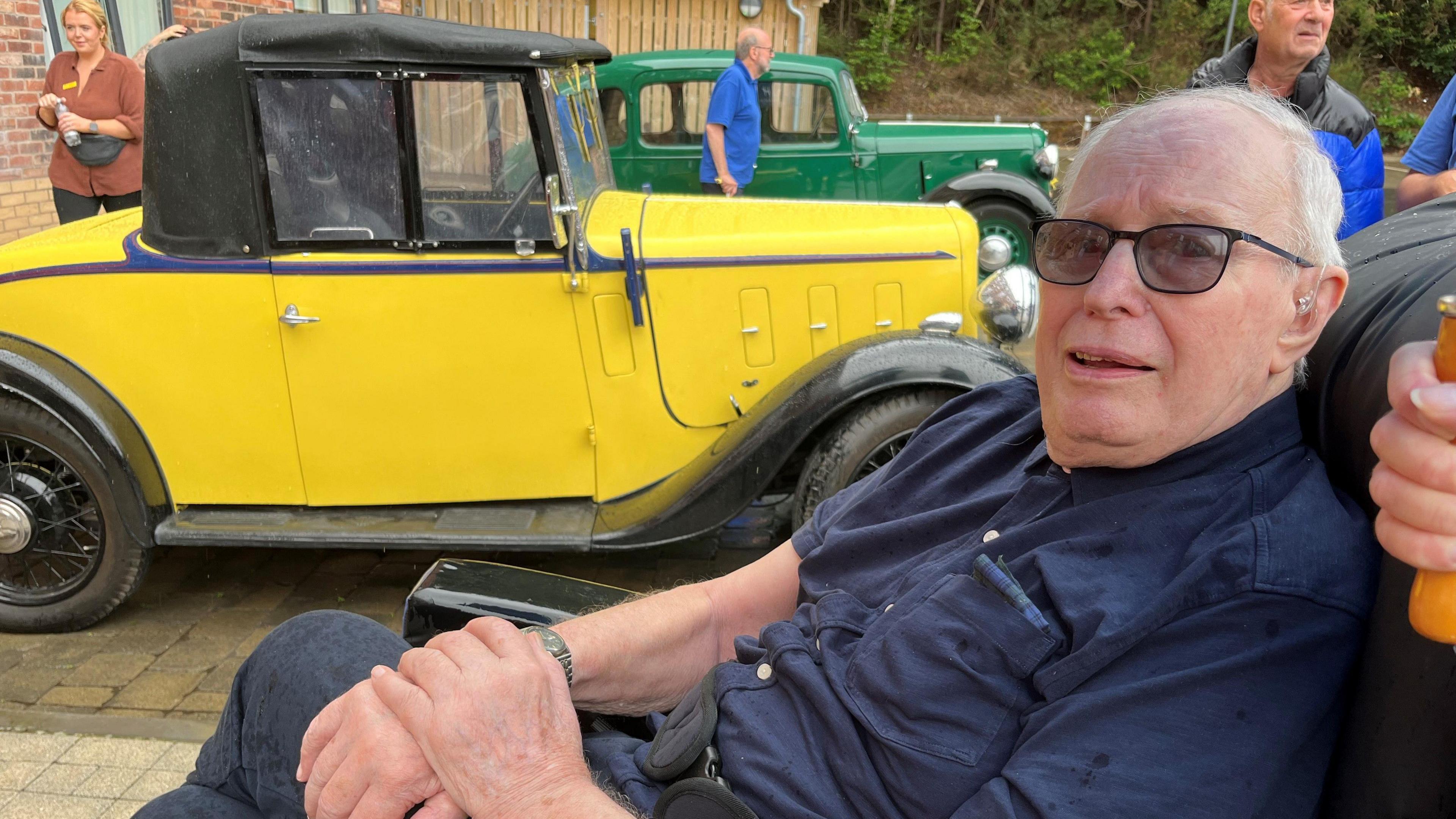 An elderly man with short white hair, glasses, a blue shirt, with his hands clasped, sits in a wheelchair and looks at the camera. Behind him is an old yellow car with a black roof. 