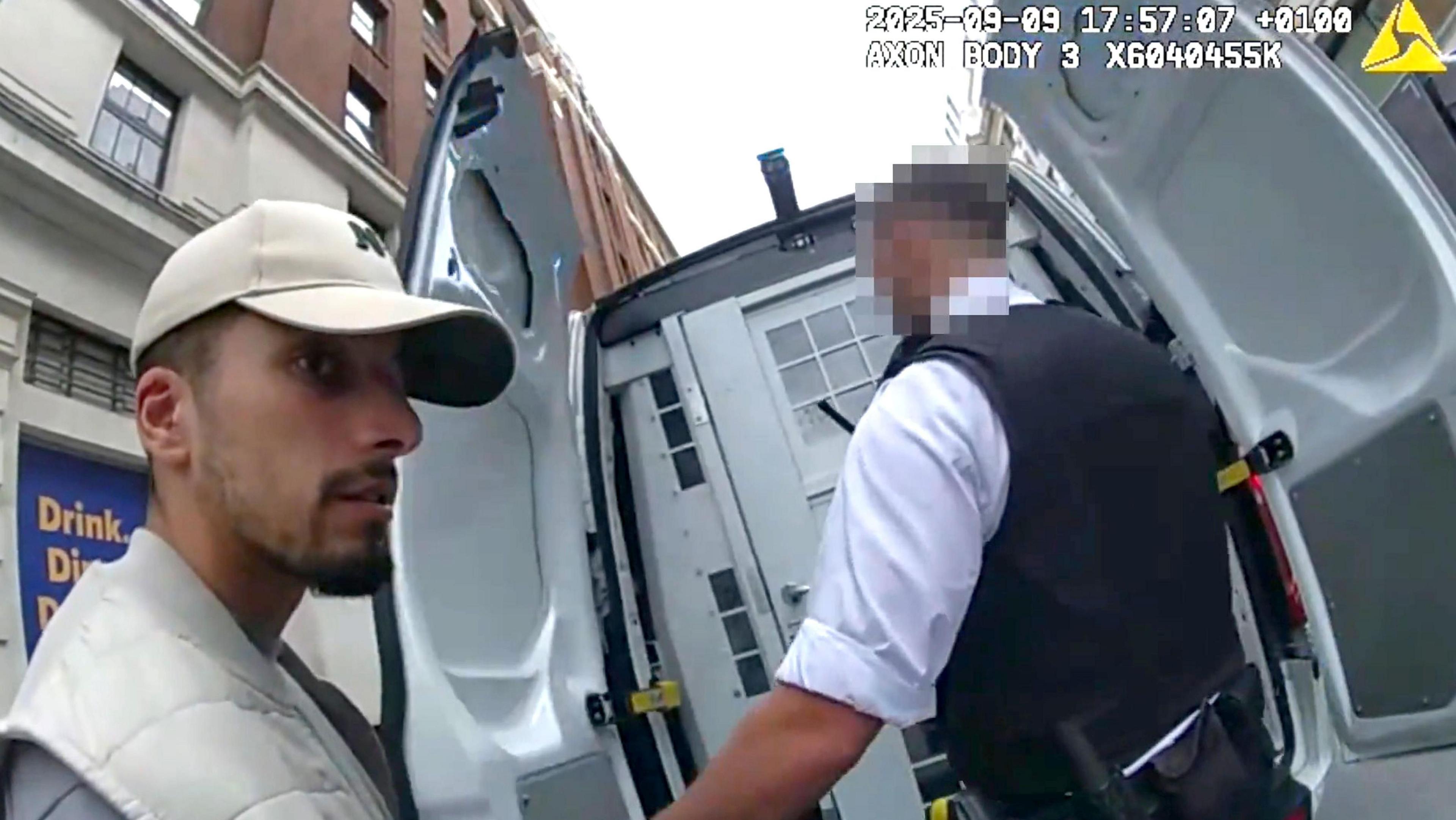 Brahim Kaddour-Cherif seen in arrest footage from police bodycam. He is wearing a baseball cap and white sleeveless coat with a grey top on underneath. He is being led into a police van by an officer wearing body armour.