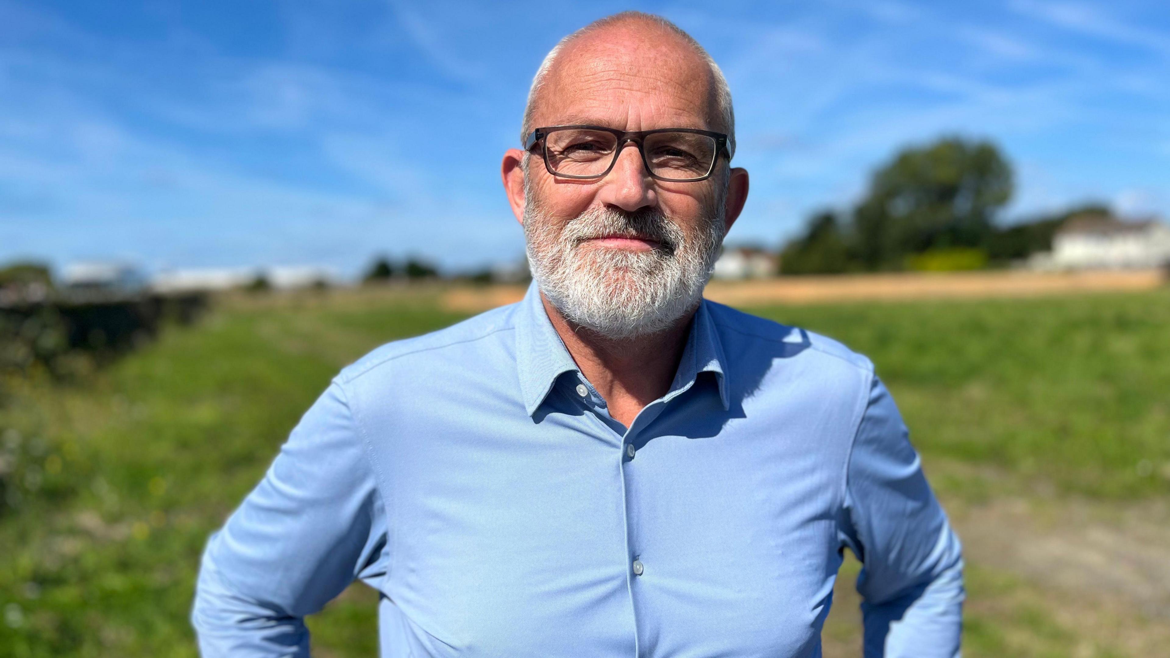 A man with a grey beard, blue shirt and black glasses smiling with his hands on his hips. 