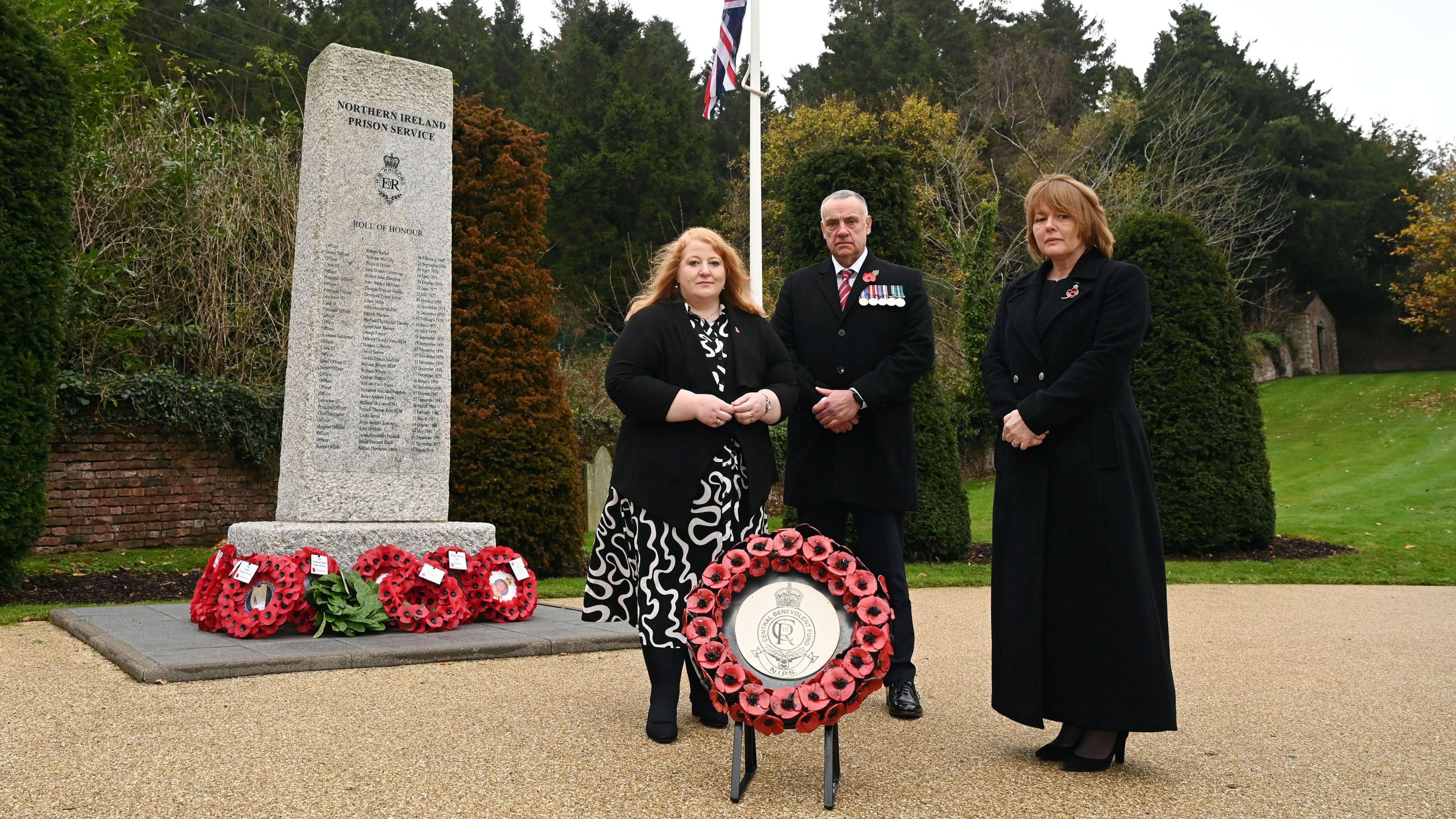 Naomi Long standing at a cenotaph wearing black alongside two others who are also wearing black. The commemorative ceramic Poppy Wreath is sitting in front of them.