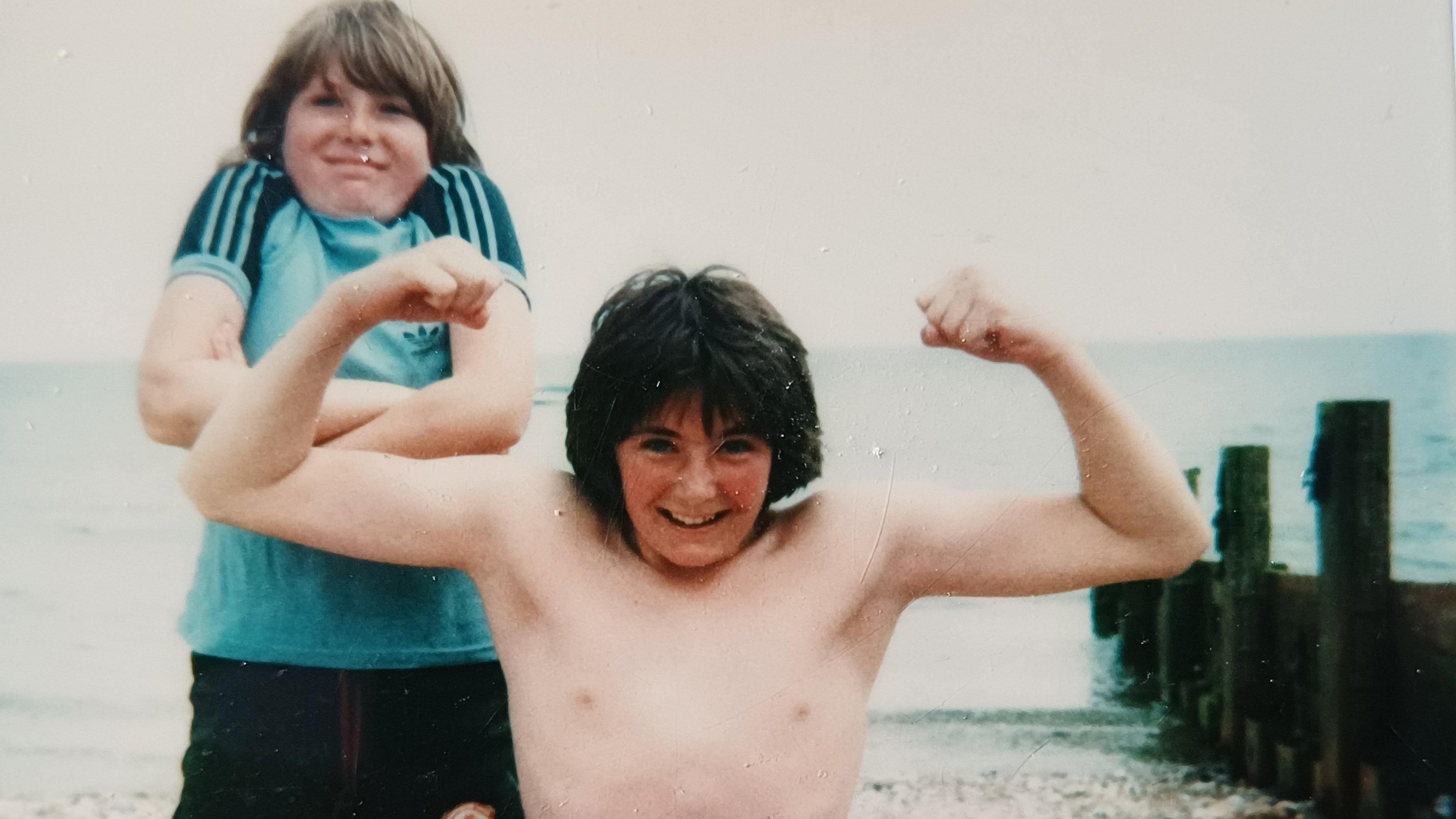 Teenager Nicky shrugs his shoulders behind his older brother Patrick who flexes his muscles while on a family holiday on a beach in the UK   