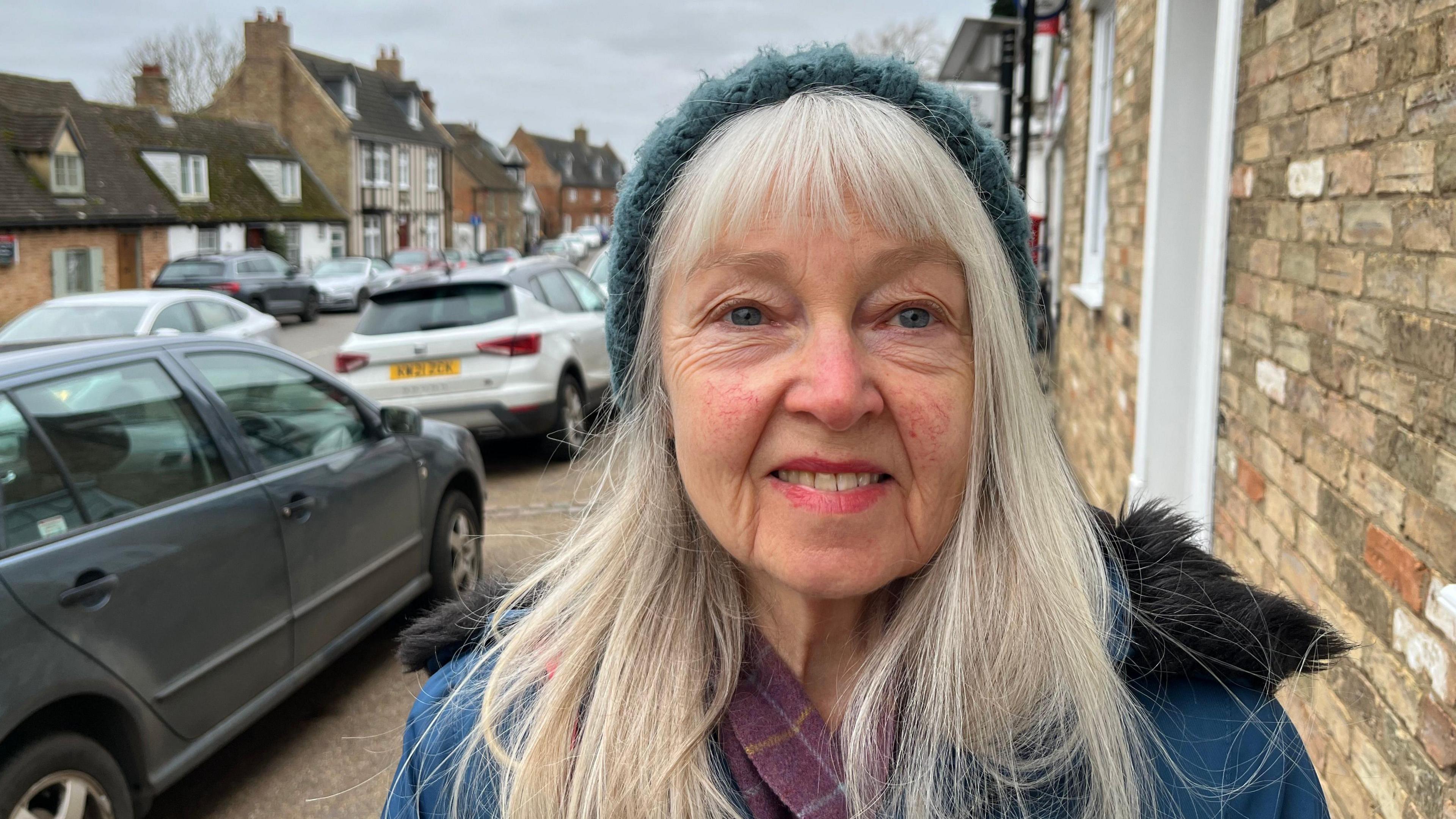 Alice Parr smiles at the camera as she is photographed on a street. She is wearing a blue coat and grey woolly hat. She has long white hair. There are a number of cars parked along the street.  