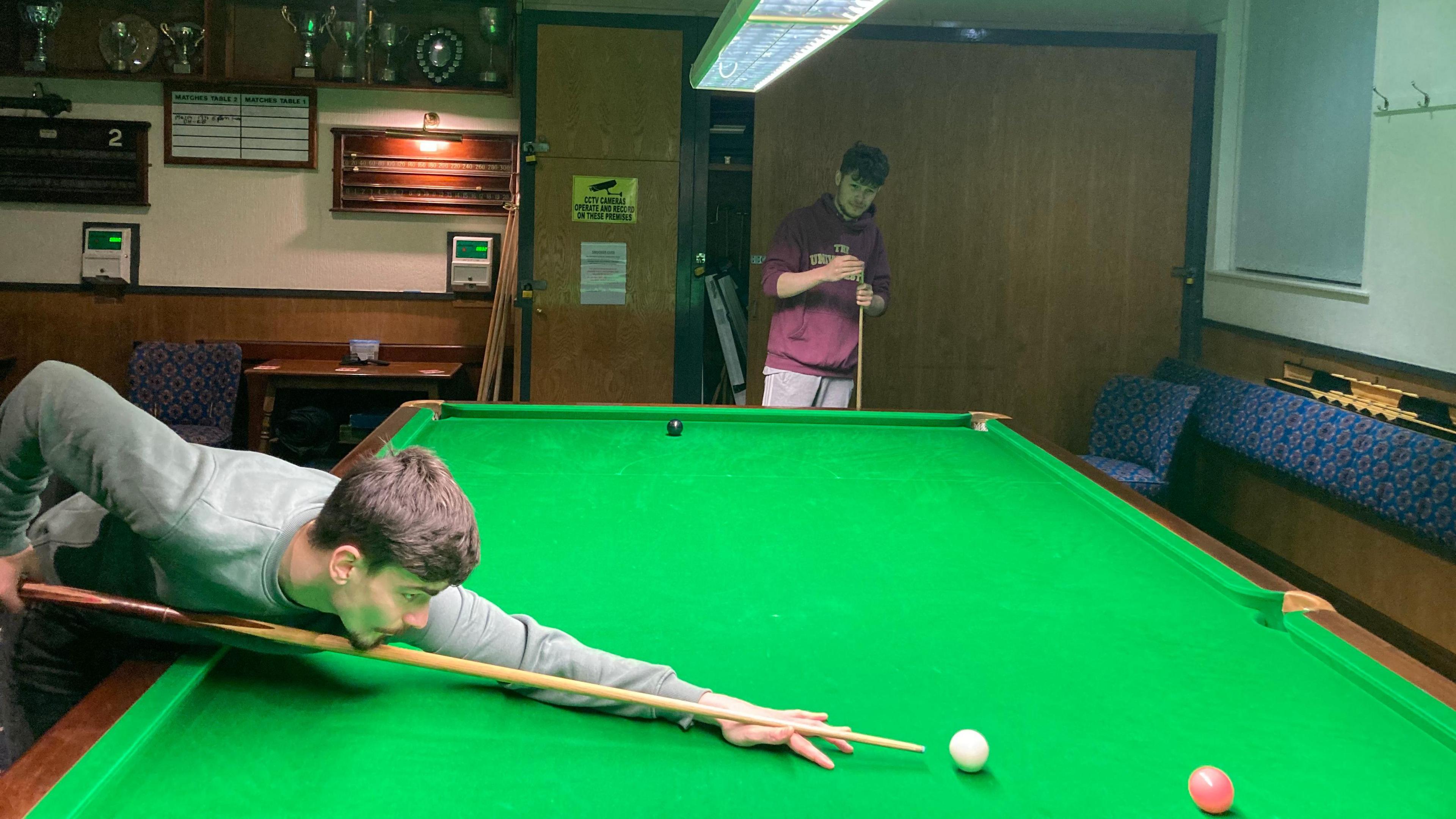 Two young men play snooker at a full-sized table within a social club. One is leaning across the baize with his chin on his cue, preparing to strike the white ball towards the pink ball. The other is standing at the head of the table chalking his cue.