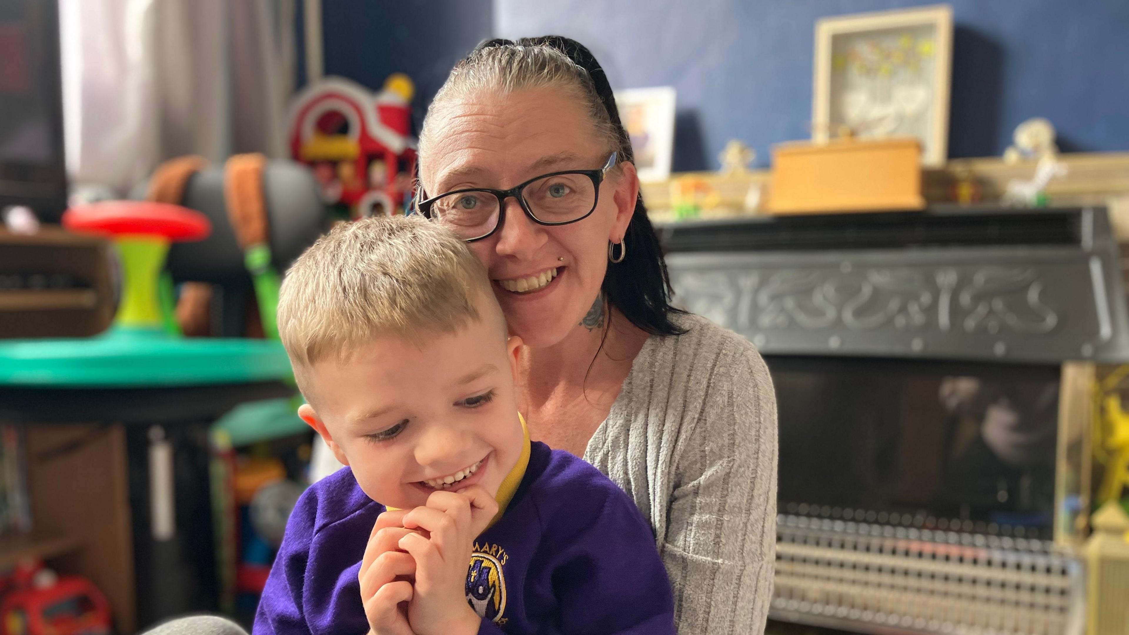 A woman in a grey top with long dark hair and glasses sits with her five year old son on her knee. They are both smiling.