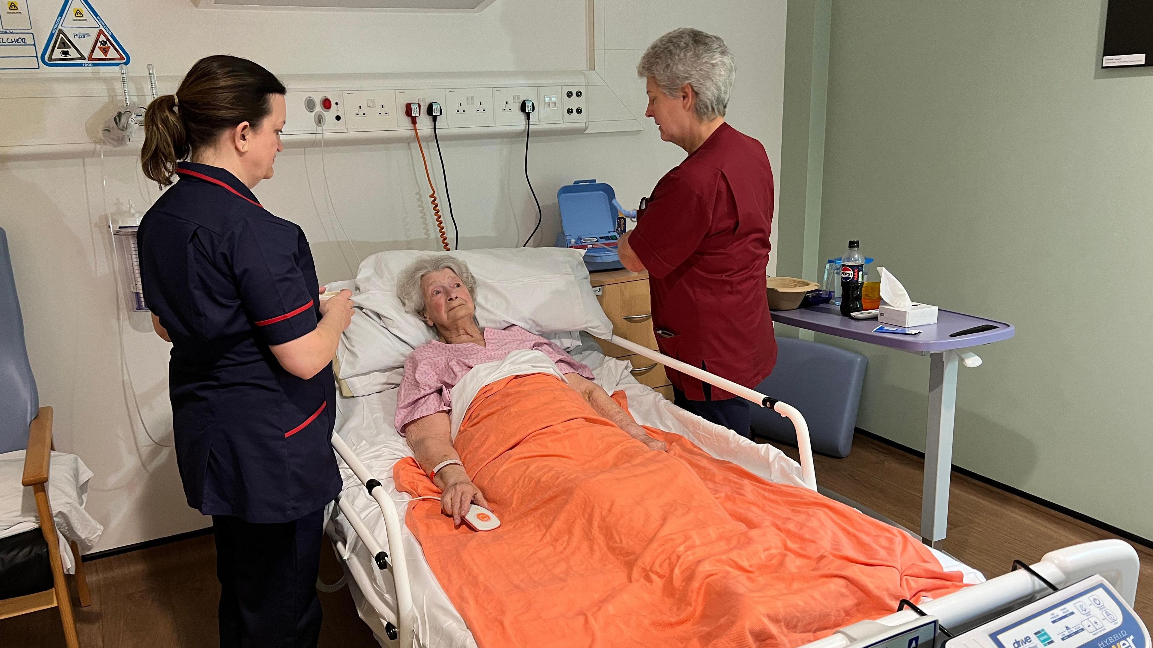 Olwen Belcher in a hospital bed, with two medical professionals tending to her on either side