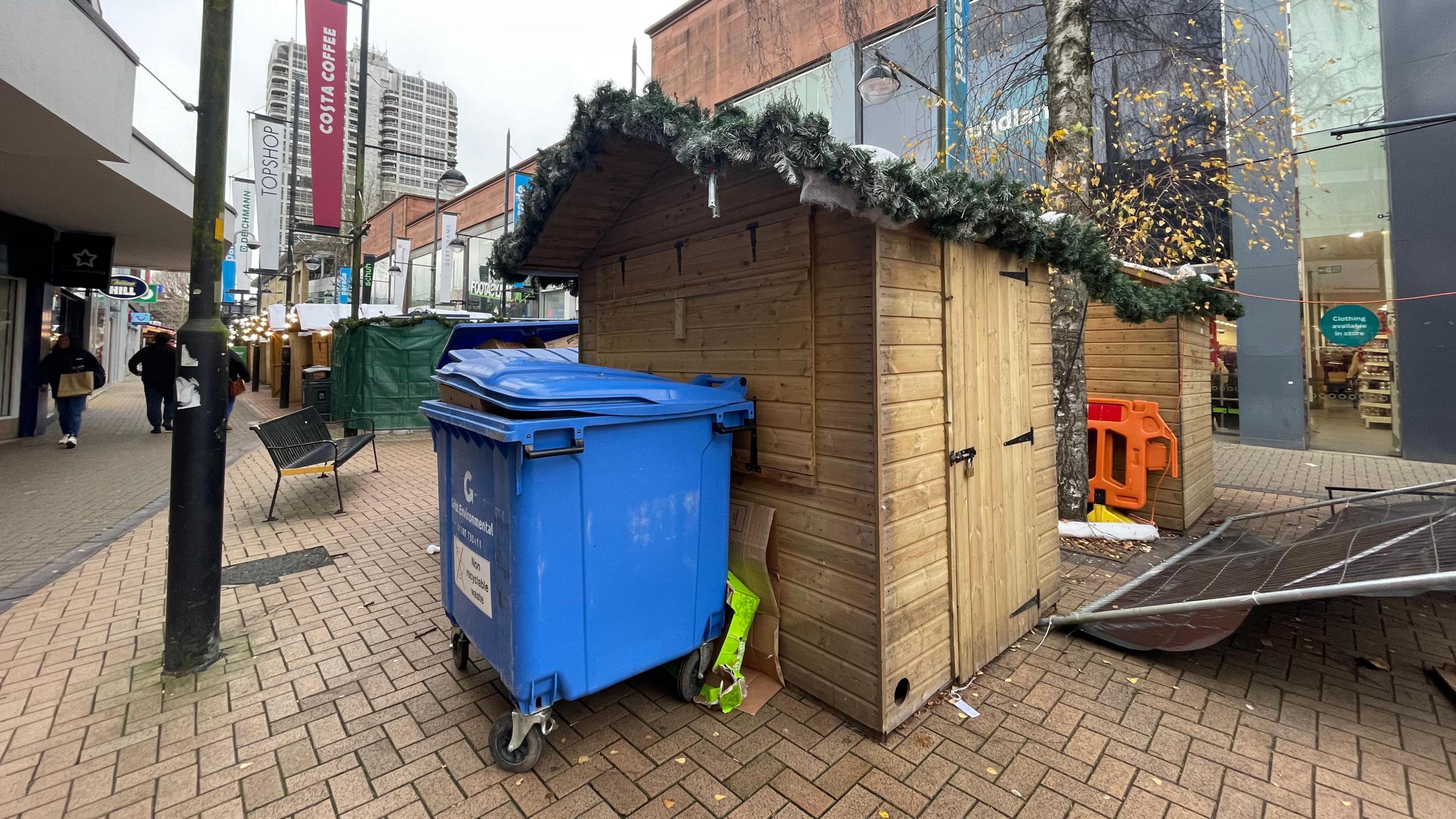 A large blue wheelie bin stands in front of a closed wooden stall at Swindon Christmas market