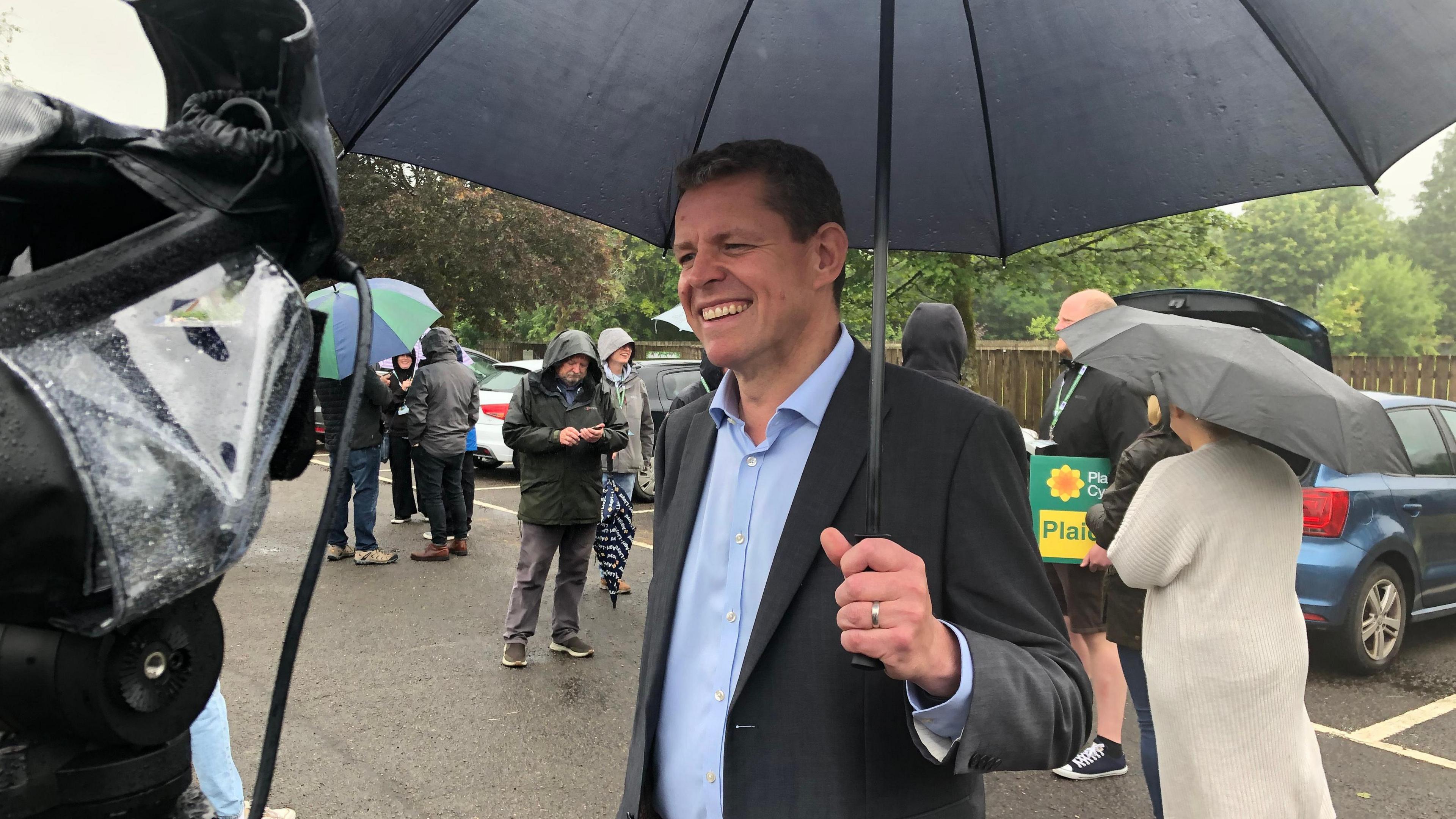 A smiling Rhun ap Iorwerth holding an umbrella in a car park