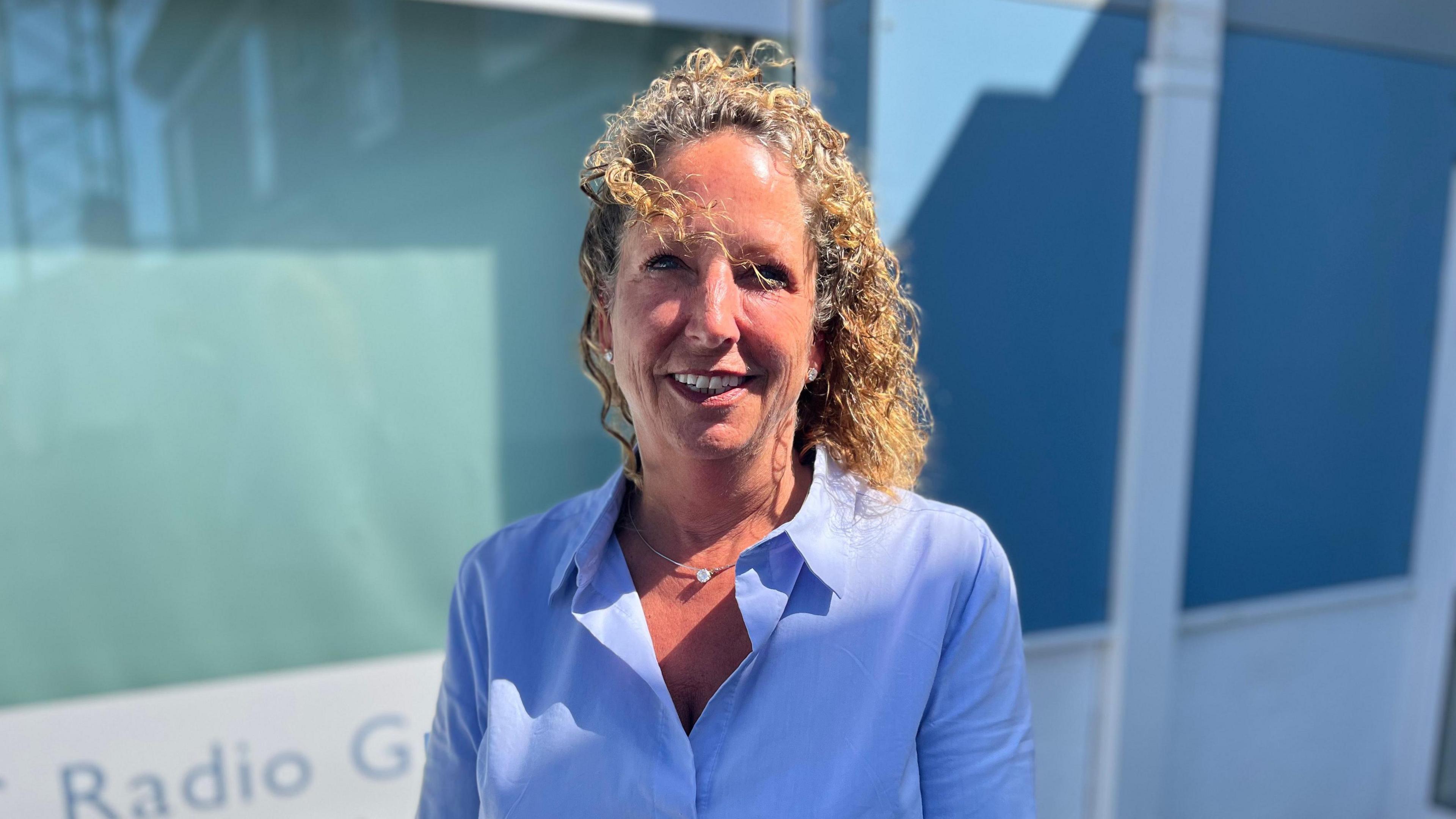 Paula Burbridge, with curly blonde hair and and in a blue blouse, smiling at the camera, standing outside BBC Radio Guernsey