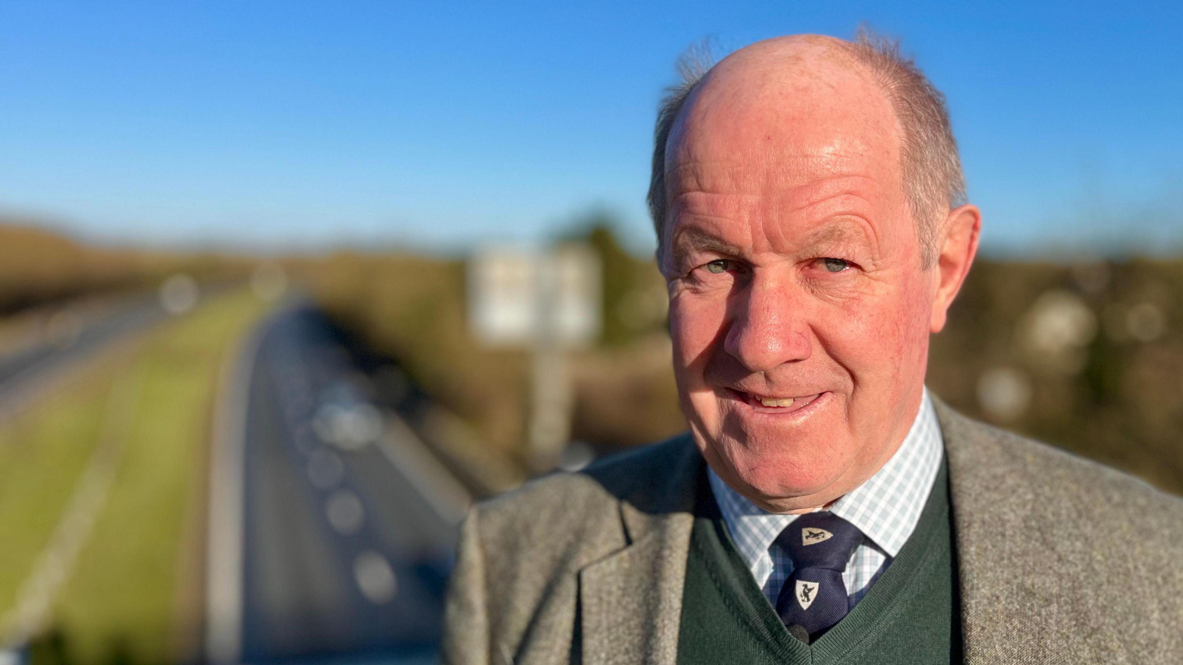 Tim Passmore smiles at the camera while standing on a bridge over the A14 carriageway. He is largely bald with some grey hair. He wears a grey suit jacket with a green jumper, shirt and tie underneath.