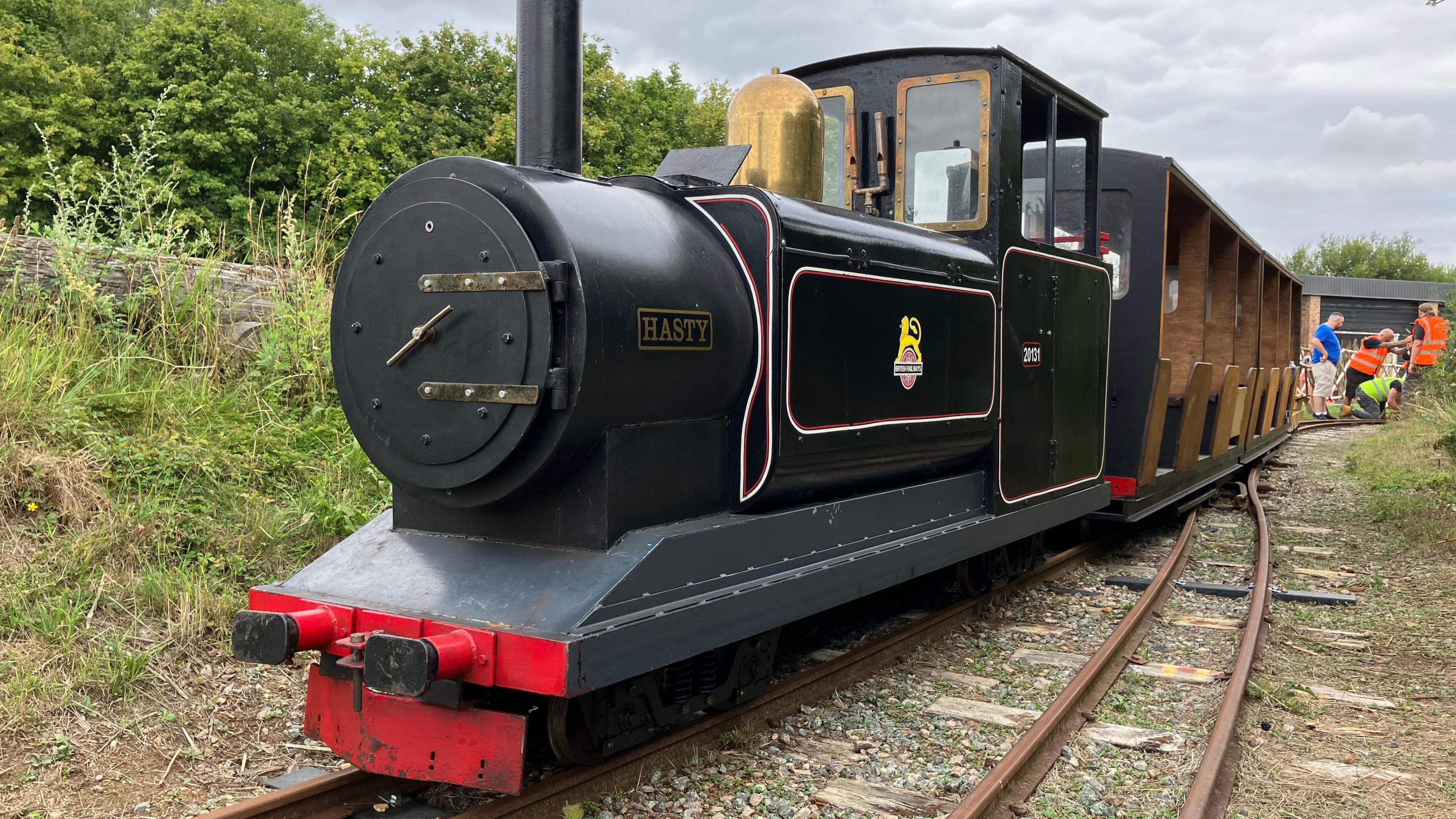 A small black locomotive called Rusty with two open sided passenger carriages wait for people to carry out work on the track.