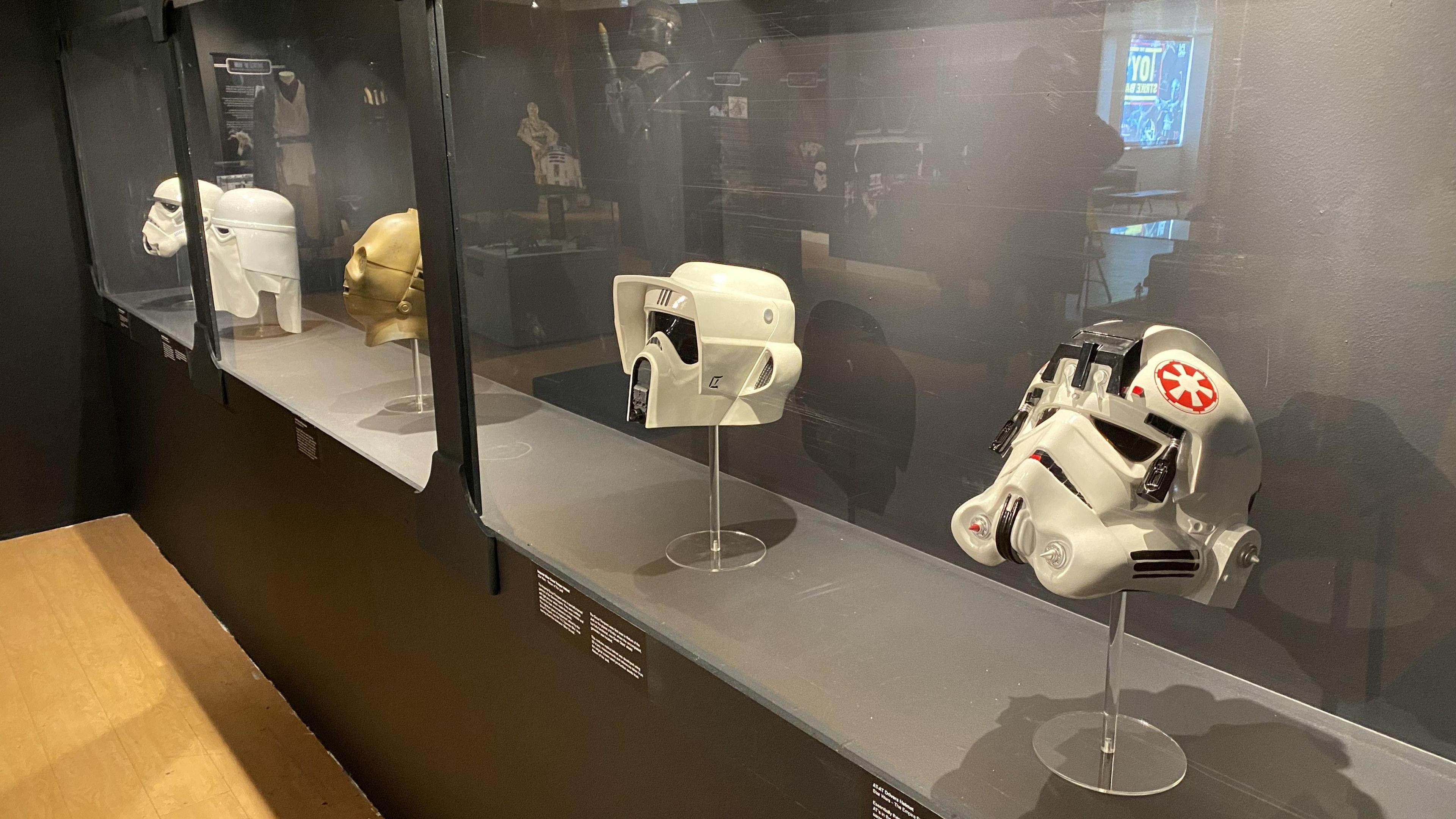 Five helmets in a glass case. They include a white Stormtrooper mask and the gold mask of droid C3PO. There are also three variants of the Stormtrooper helmet as appearing in the films. They have curved bottom halves, and more angular tops around the forehead area. There are eyes in the shape of dark lenses, and in the centre of the helmets, around the mouth area of a wearer, there are grills in the shape of an arrow head, which makes it look like the helmets are frowning.