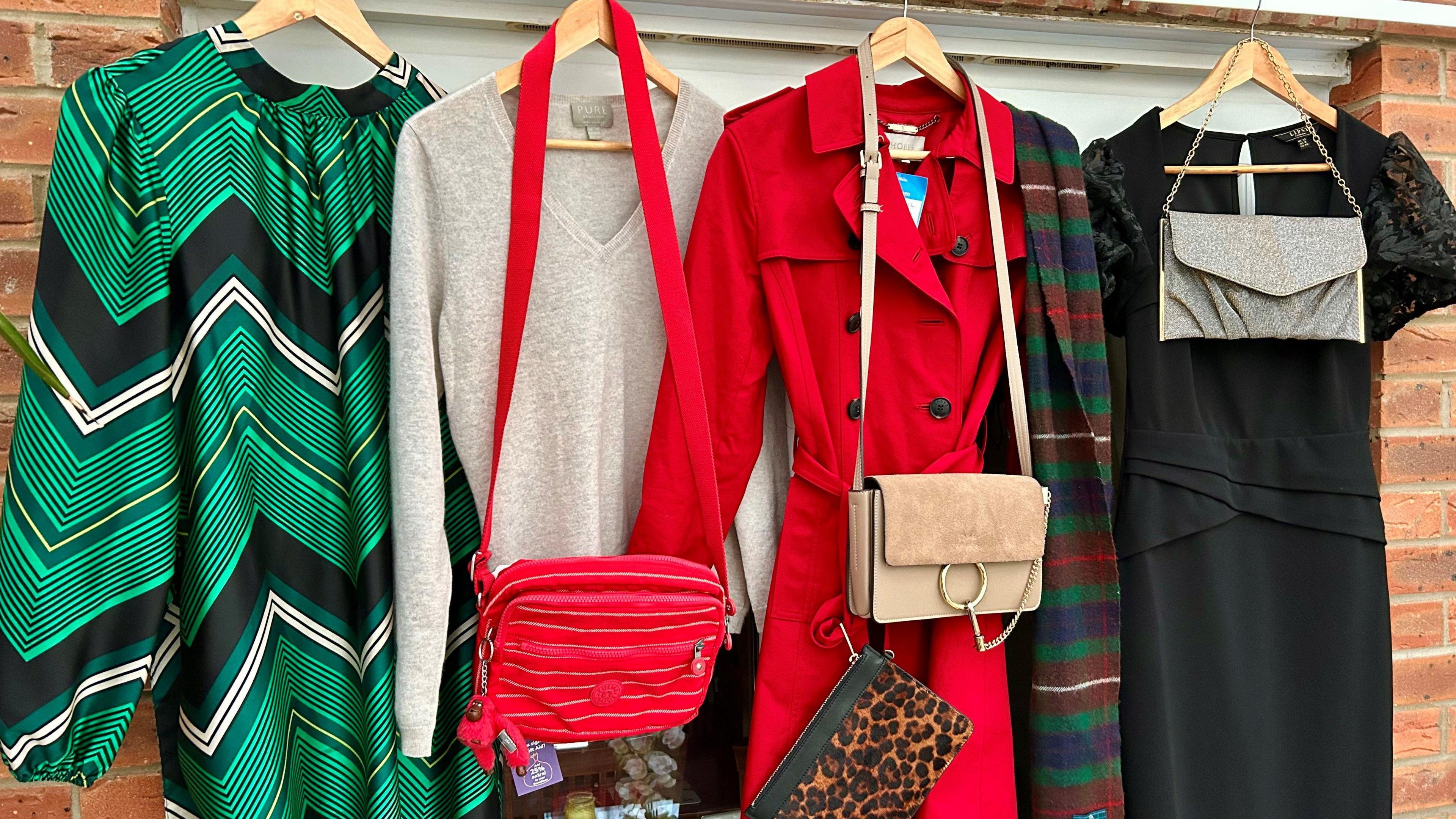 Four items of clothing hanging up in a room on hangers. A green pattern dress, a grey jumper, with a red bag over it, a red coat, a leopard print bag, a cream bag, a tartan scarf, a black dress and a grey handbag, are all in front of a wall. 