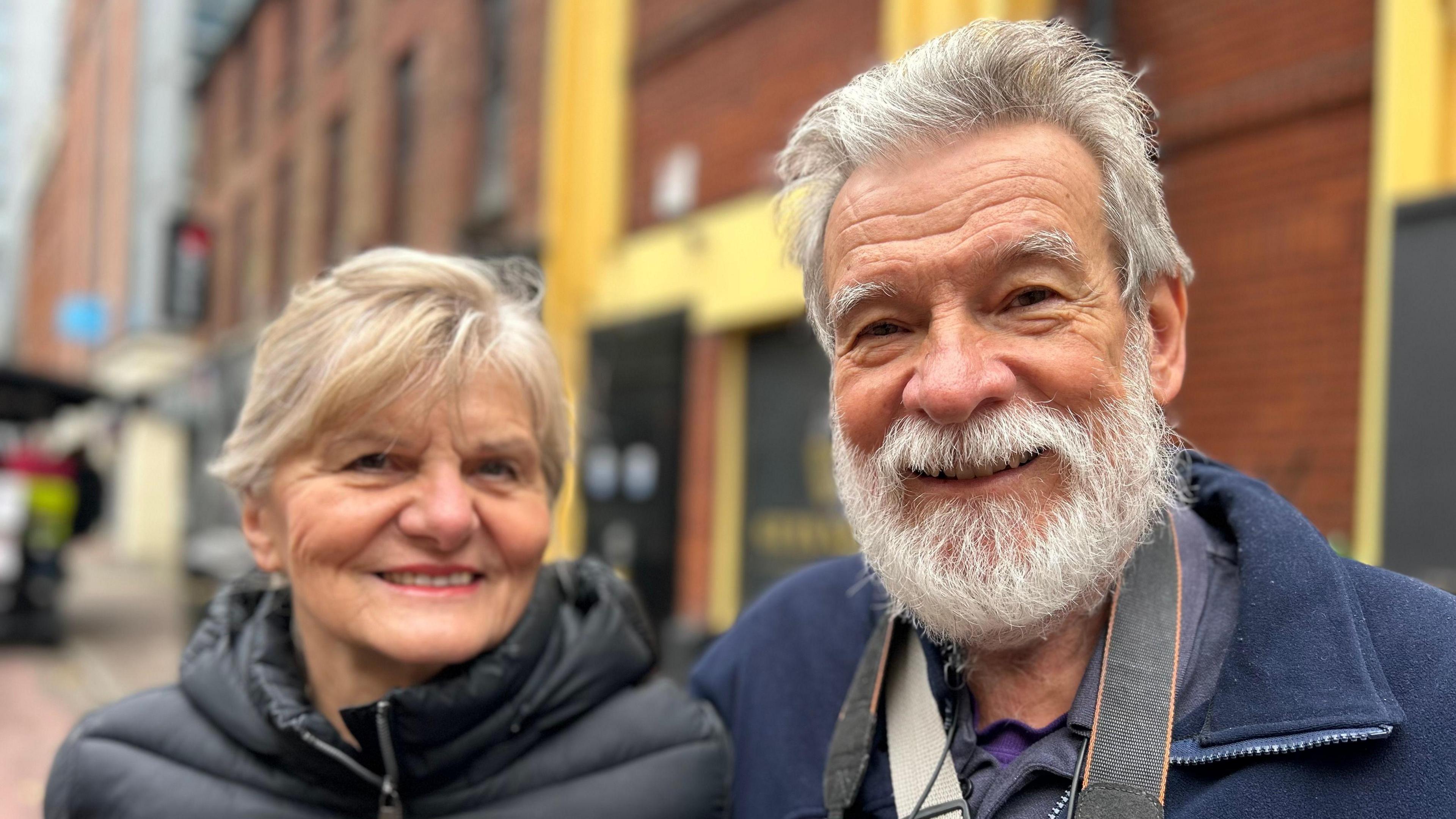 Monica and John Frim smiling while in Belfast. Monica is wearing a black puffer coat and John is wearing a navy fleece with a camera strap around his neck