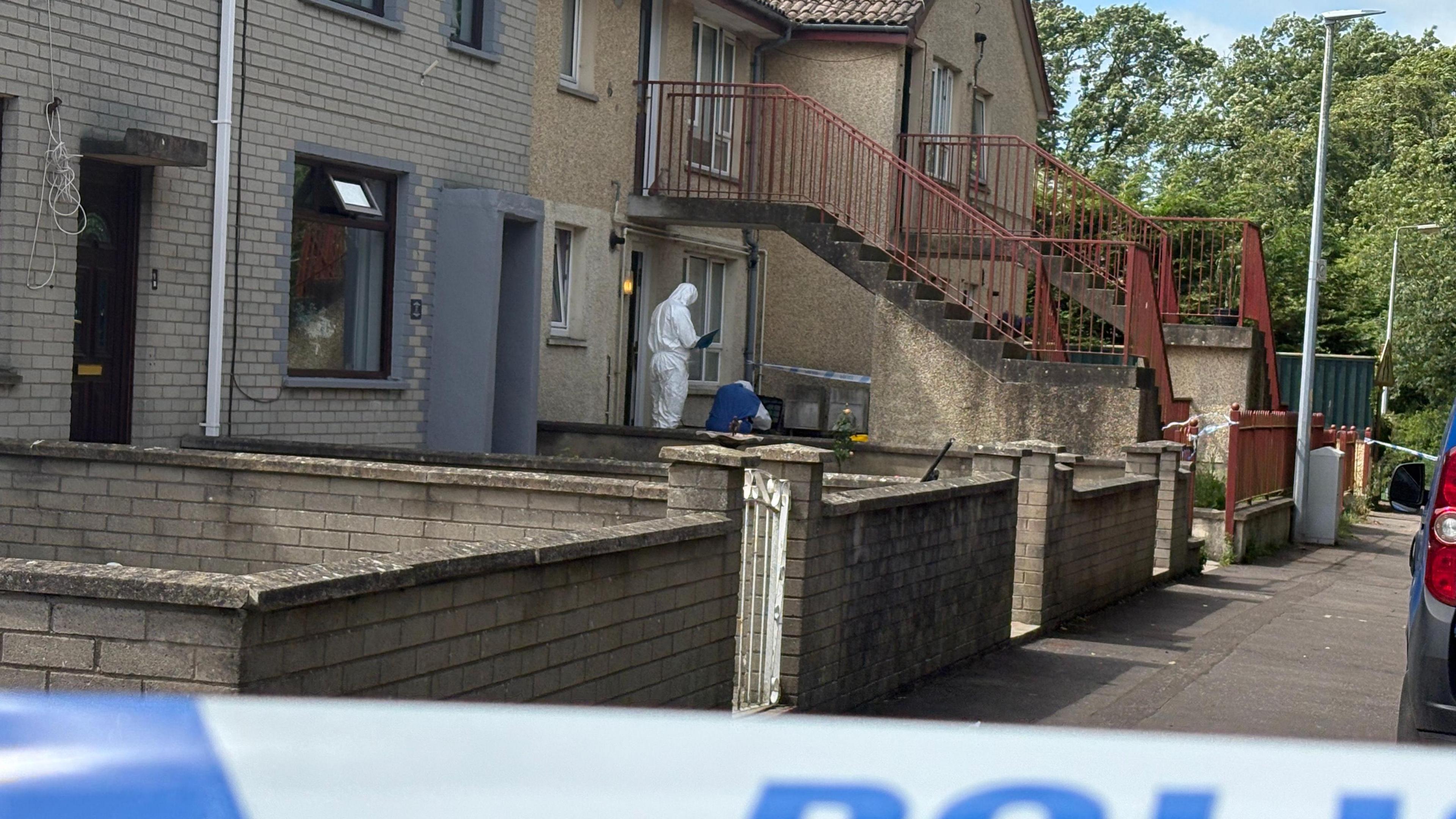 A forensic investigator standing outside a row of terraced properties at Elmfield Walk in Donaghadee where Sarah Montgomery died on Saturday.  The investigator is dressed in a white hazmat suit and is holding a clipboard.  The properties are two stories in height and there are a couple of sets of concrete steps leading to the first floor properties.  There are cordons to the front and side of one ground floor property. 