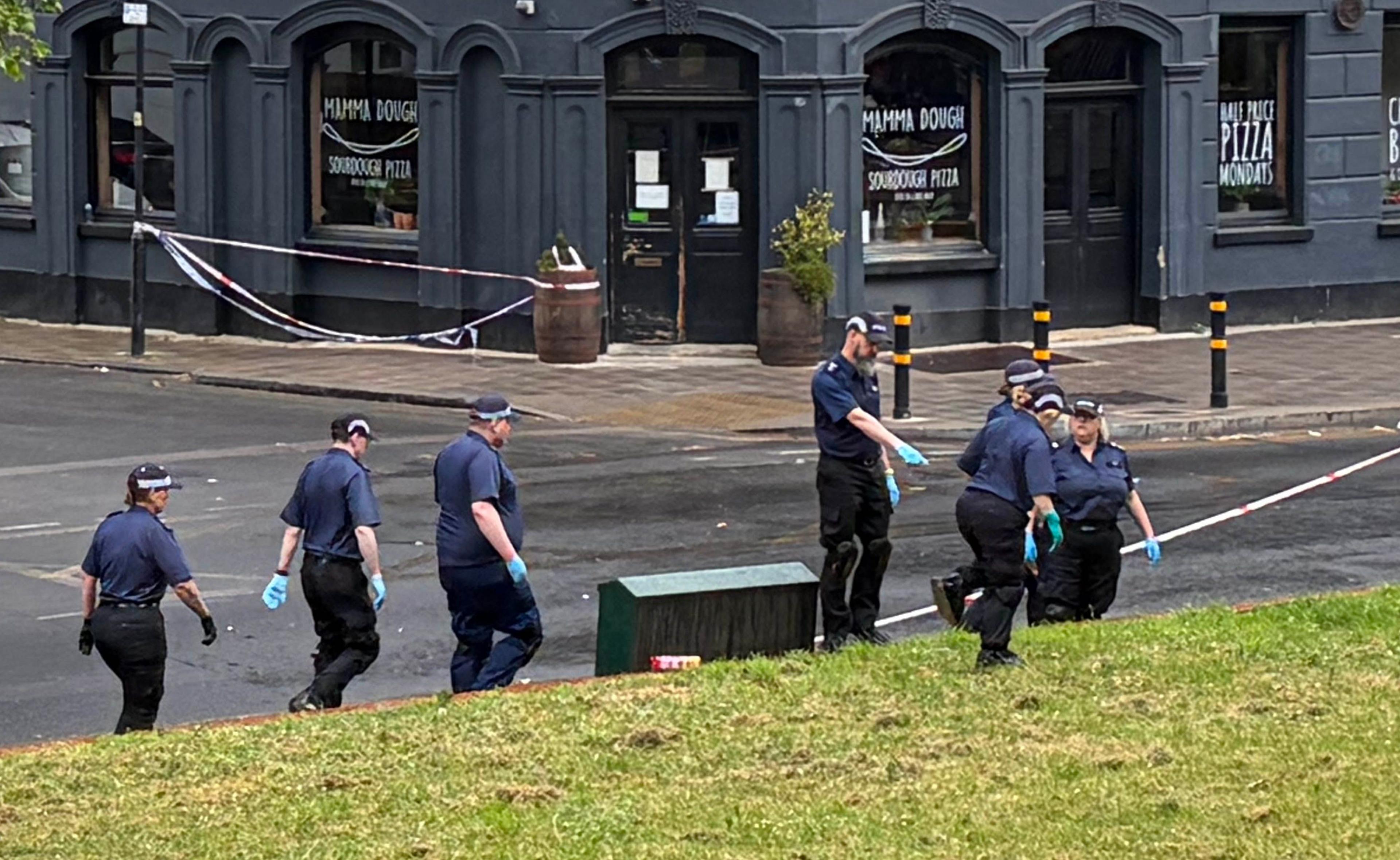 Shtatë oficerë policie me bluza blu me mëngë të shkurtra, kapele të zeza dhe doreza kirurgjikale blu kontrollojnë një zonë me bar. Në sfond, shihet fasada e një restoranti gri me mbishkrimin "Mamma Dough Sourdough Pizza" në dritare.