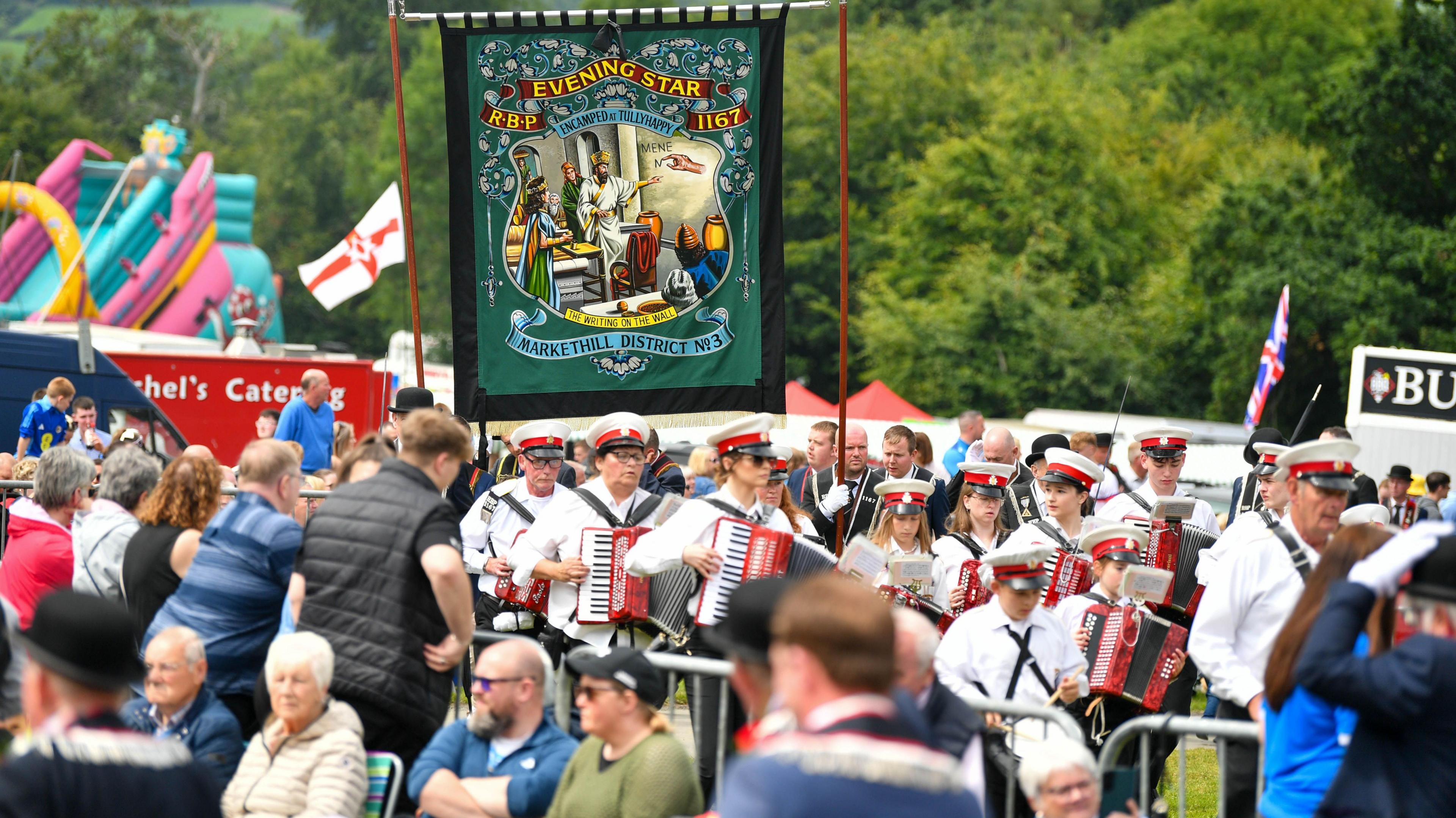 A wide shot of a band playing accordions marching in the middle of the route in Scarva