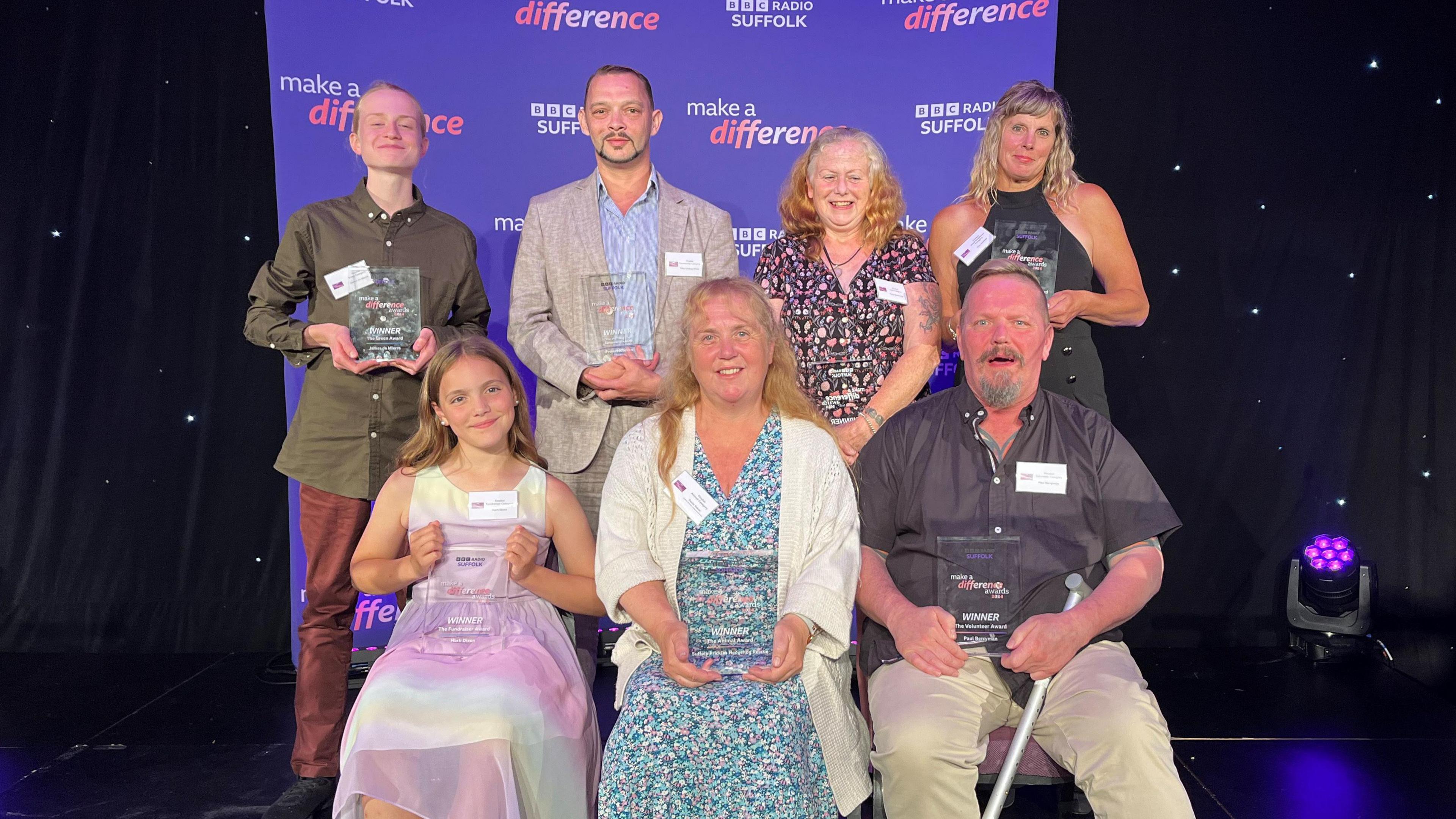 Left to right: James de Mierre, Toby Lindsay-White, Kathy Cummings, Tina Ashford, with Harli Dixon, Paula Baker and Paul Berryman in the front row. they sit on a stage in front of a banner which says 'make a difference' and each hold a glass trophy