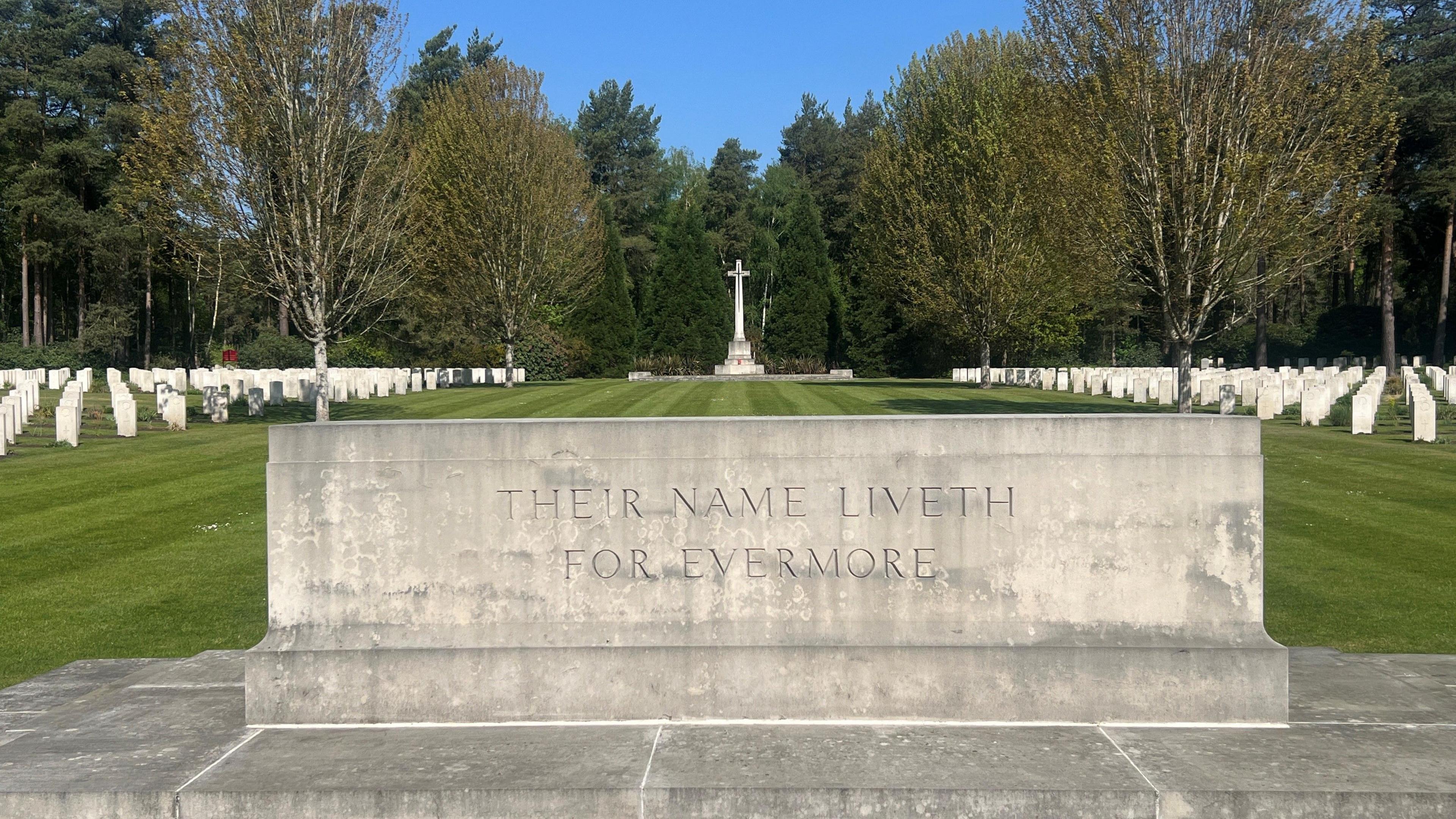 A grey stone plinth inscribed with 'Their Name Liveth For Evermore'. Dozens of white headstones in rows on neatly manicured grass and a white cross statue are seen in the background.