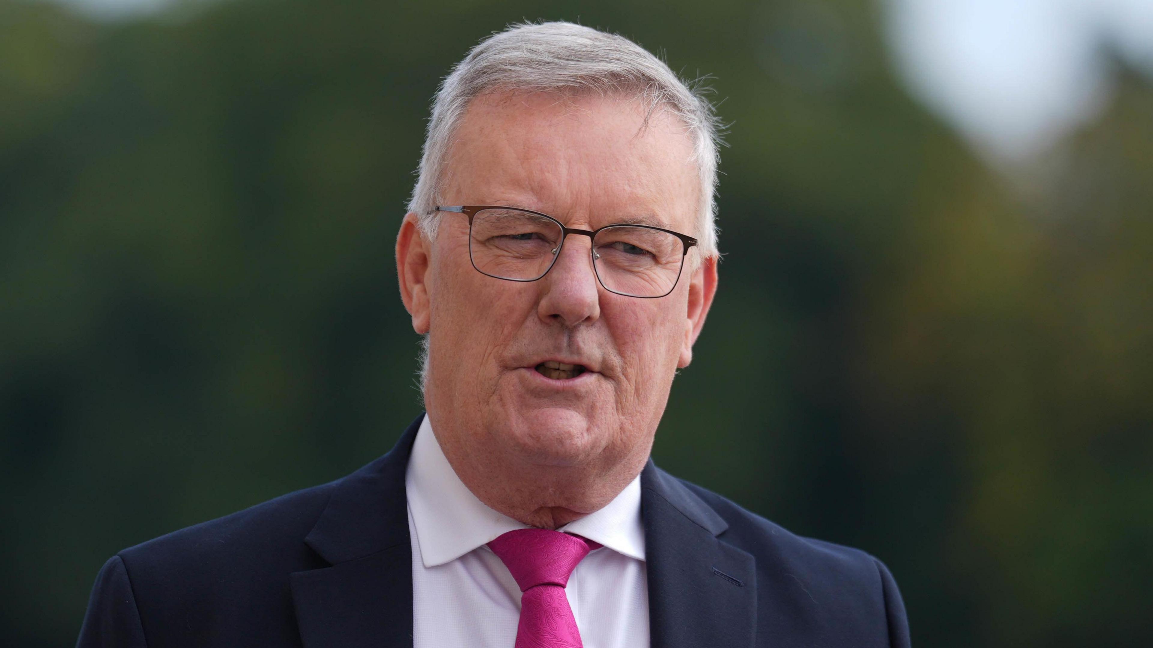 Mike Nesbitt is wearing glasses and a suit. The shirt is white with a pink tie and navy blazer. He has short white hair.