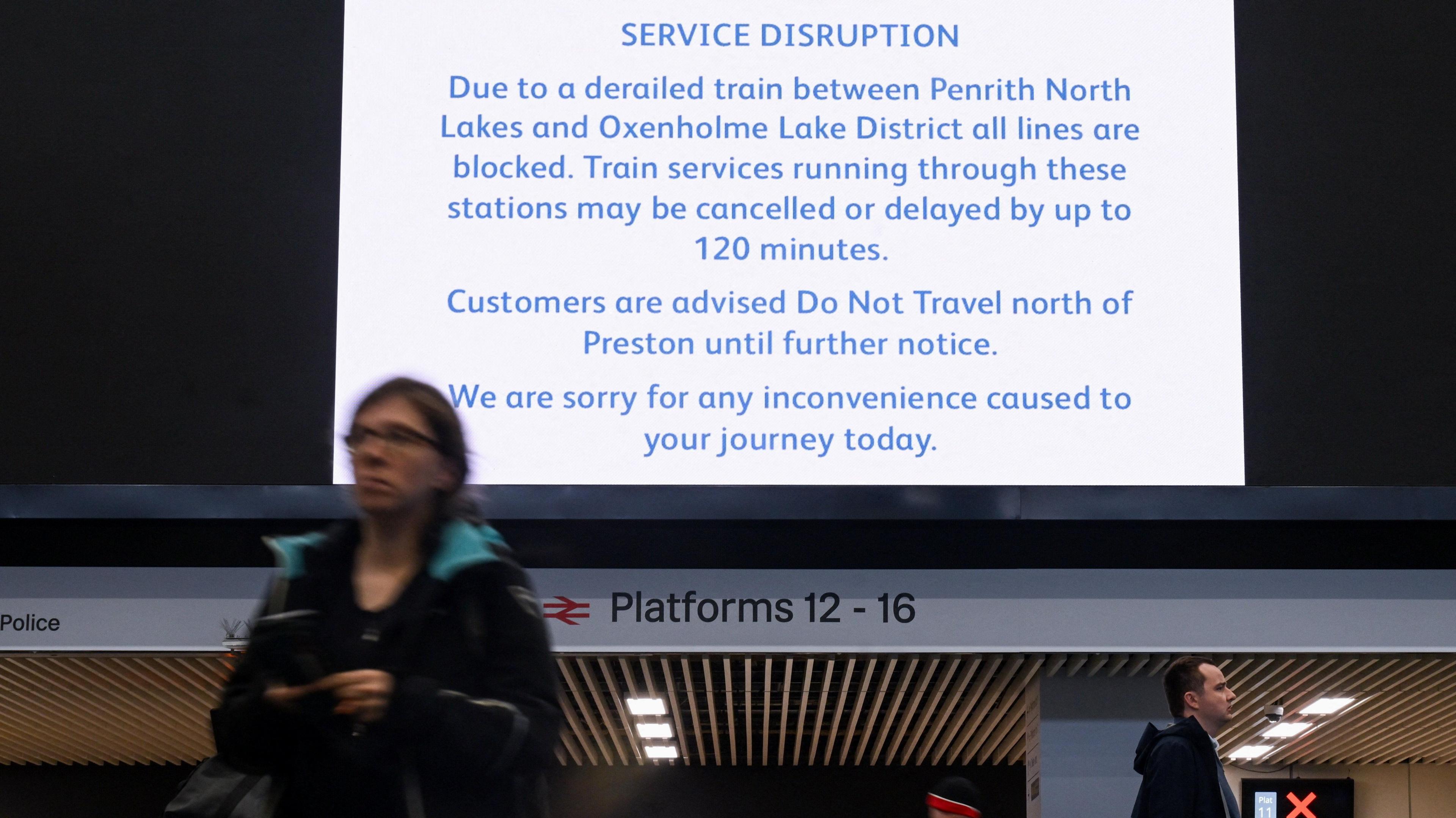 A sign at London Euston detailing the delay. There is a woman and a man walking underneath the sign. It reads 'SERVICE DISRUPTION- Due to a derailed train between Penrith North Lakes and Oxenholme Lake District all lines are blocked. Train services running through these stations may be cancelled or delayed by up to 120 minutes. Customers are advised Do Not Travel north of Preston until further notice. We are sorry for an inconvenience caused today.'