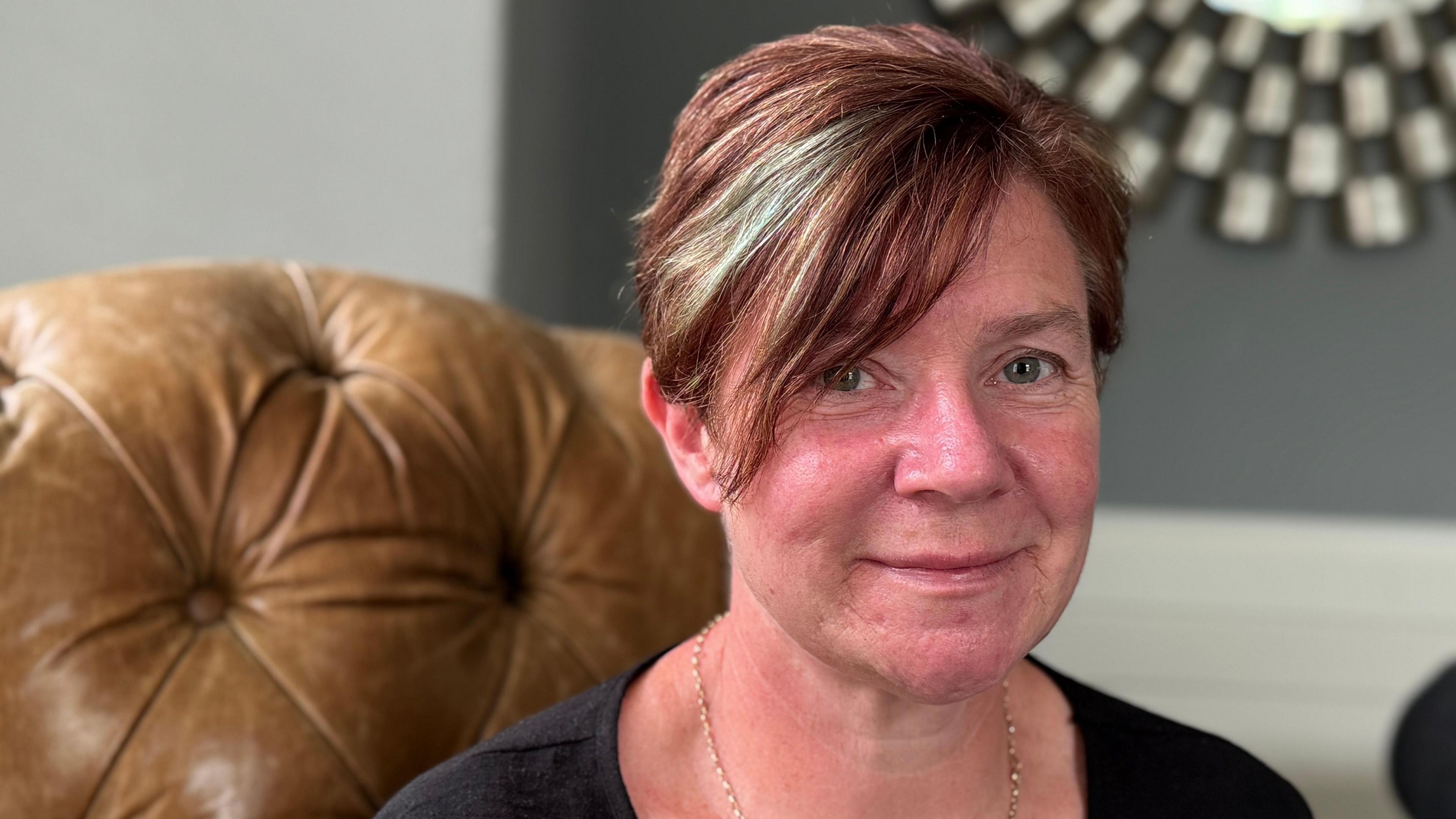 The image shows a woman sitting on a brown, tufted leather chair. They have short hair with some highlights and are wearing a black top and a thin chain necklace. In the background, there is a wall with gray paint and white trim, as well as part of a circular mirror with an intricate frame.