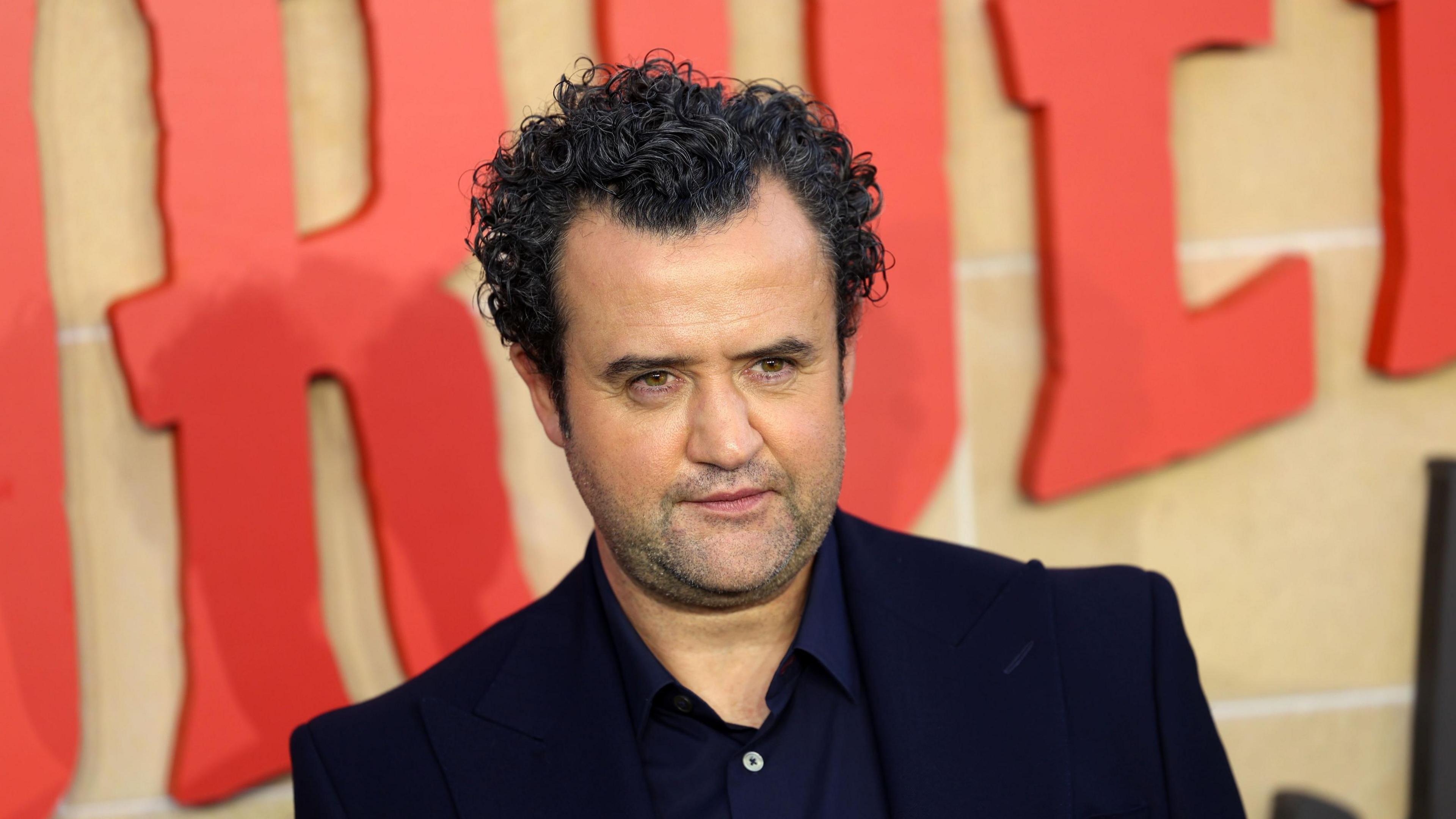 Daniel Mays is at the premier of The Thursday Murder Club in London. The title of the film is written behind him. He has olive skin and is wearing a dark blue coloured blazer and shirt.