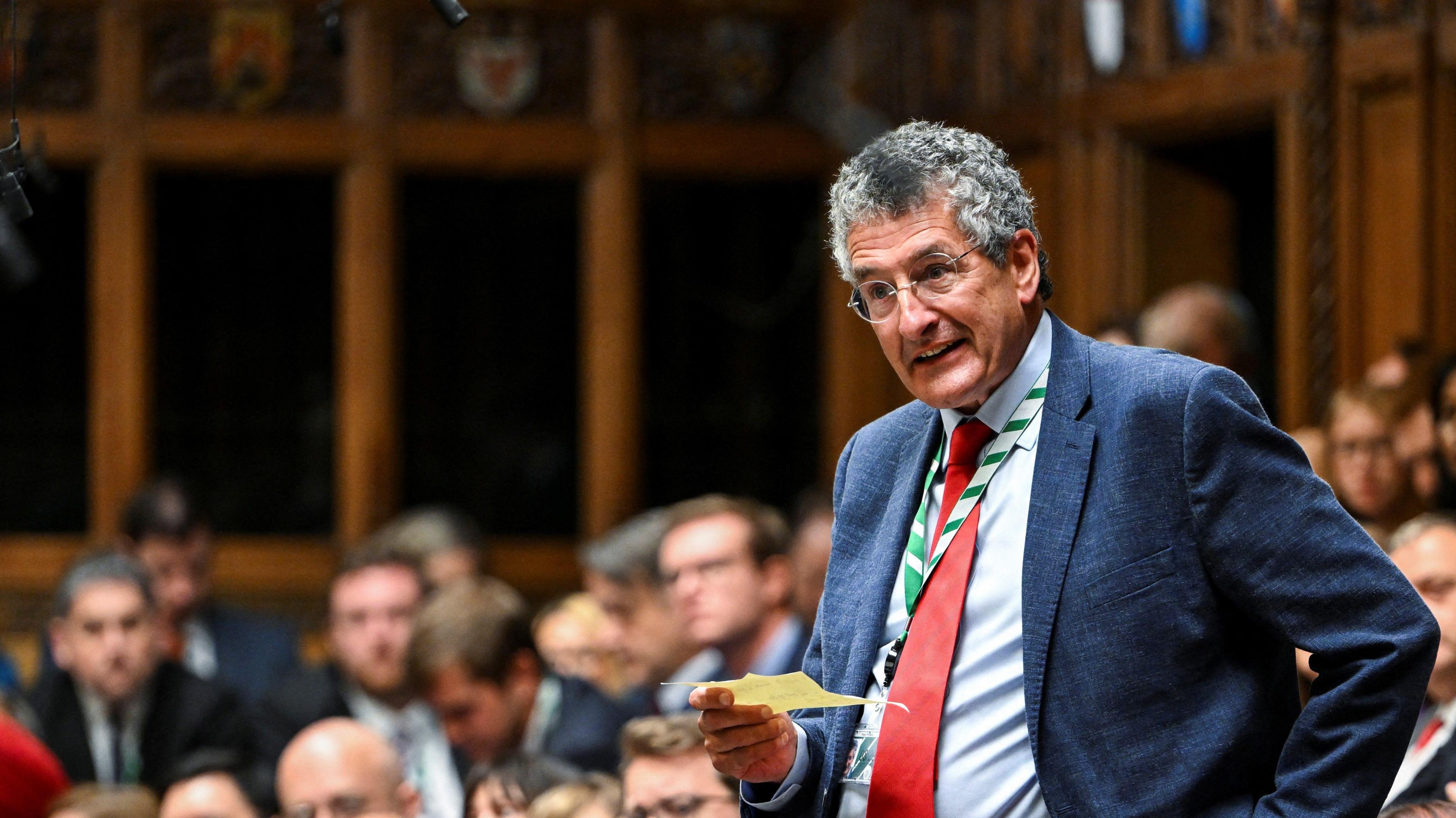 Member of Parliament Peter Prinsley speaks during Prime Minister's Questions at the House of Commons in London in September. He is pictured standing with a piece of paper in his hand. Other MPs sit around him. He is wearing a blue suit and shirt with a red tie.