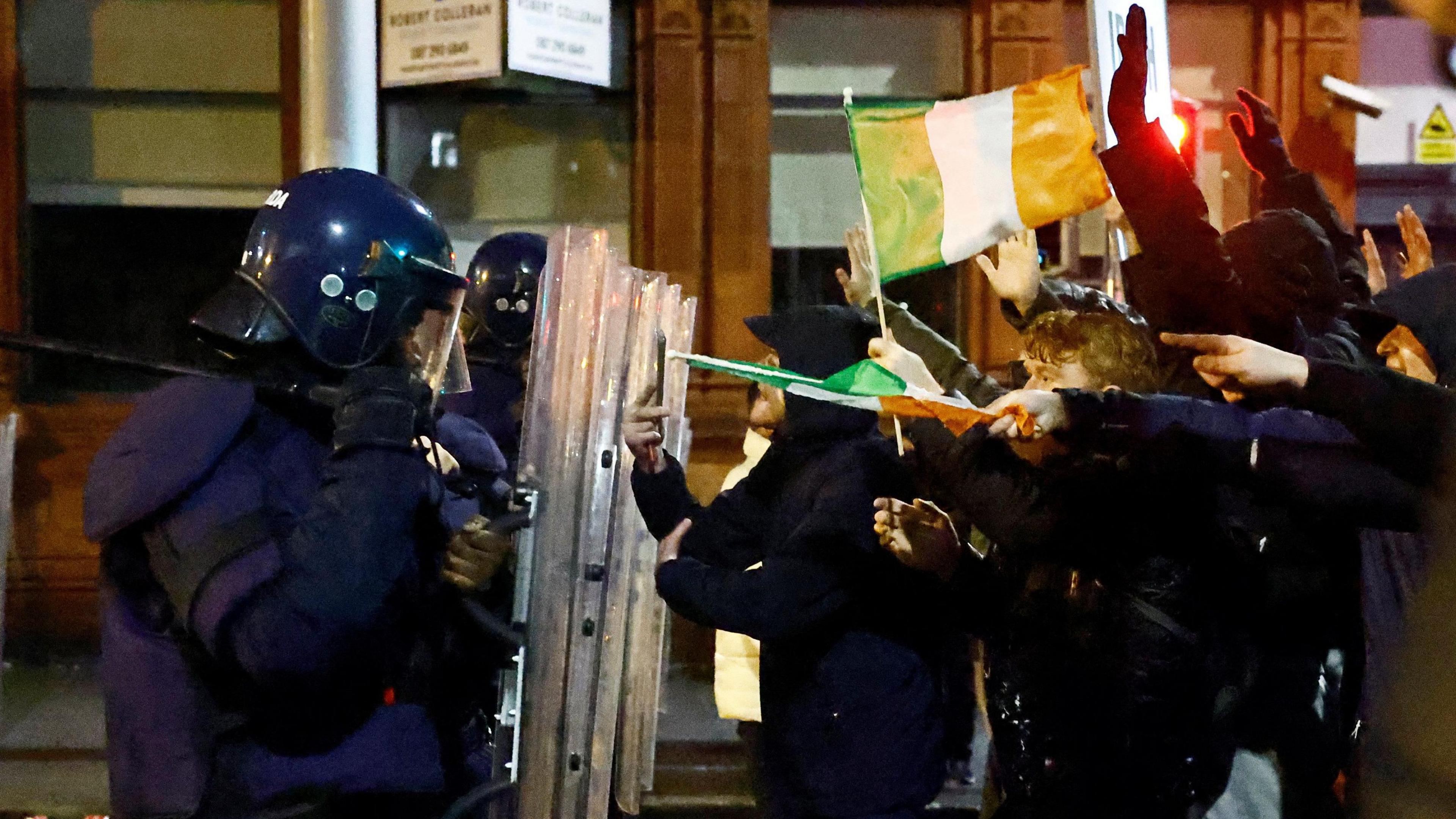 People stand in front of riot police. Protesters are holding up Irish flags while police are holding up right gear.