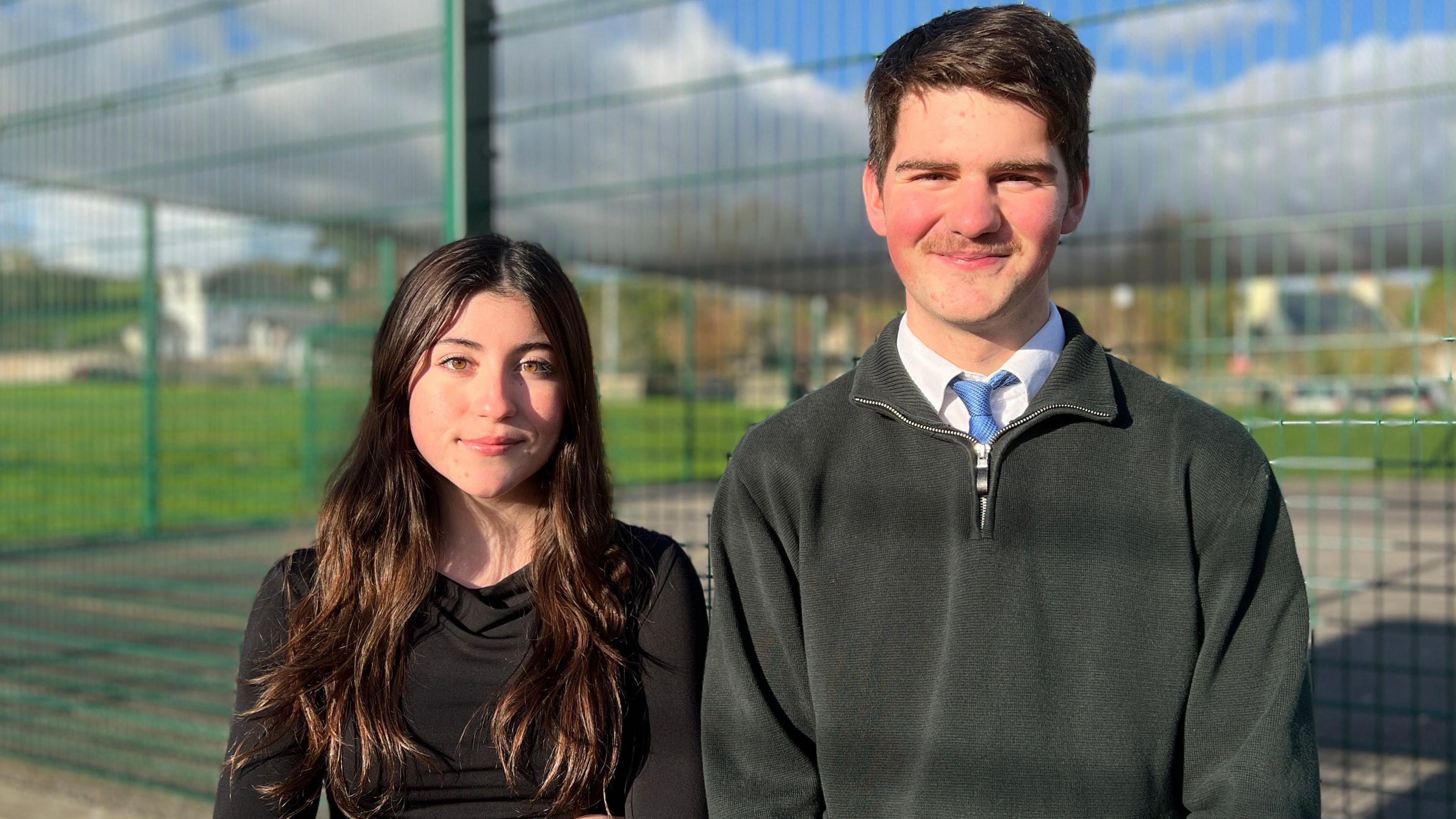 Alicia and Logan are standing outdoors in front of a tall green metal fence. The background shows a grassy area, a clear blue sky with scattered clouds, and some distant buildings. Alicia is wearing a dark long-sleeve top with arms crossed, while Logan is dressed in a dark sweater over a collared shirt and tie.