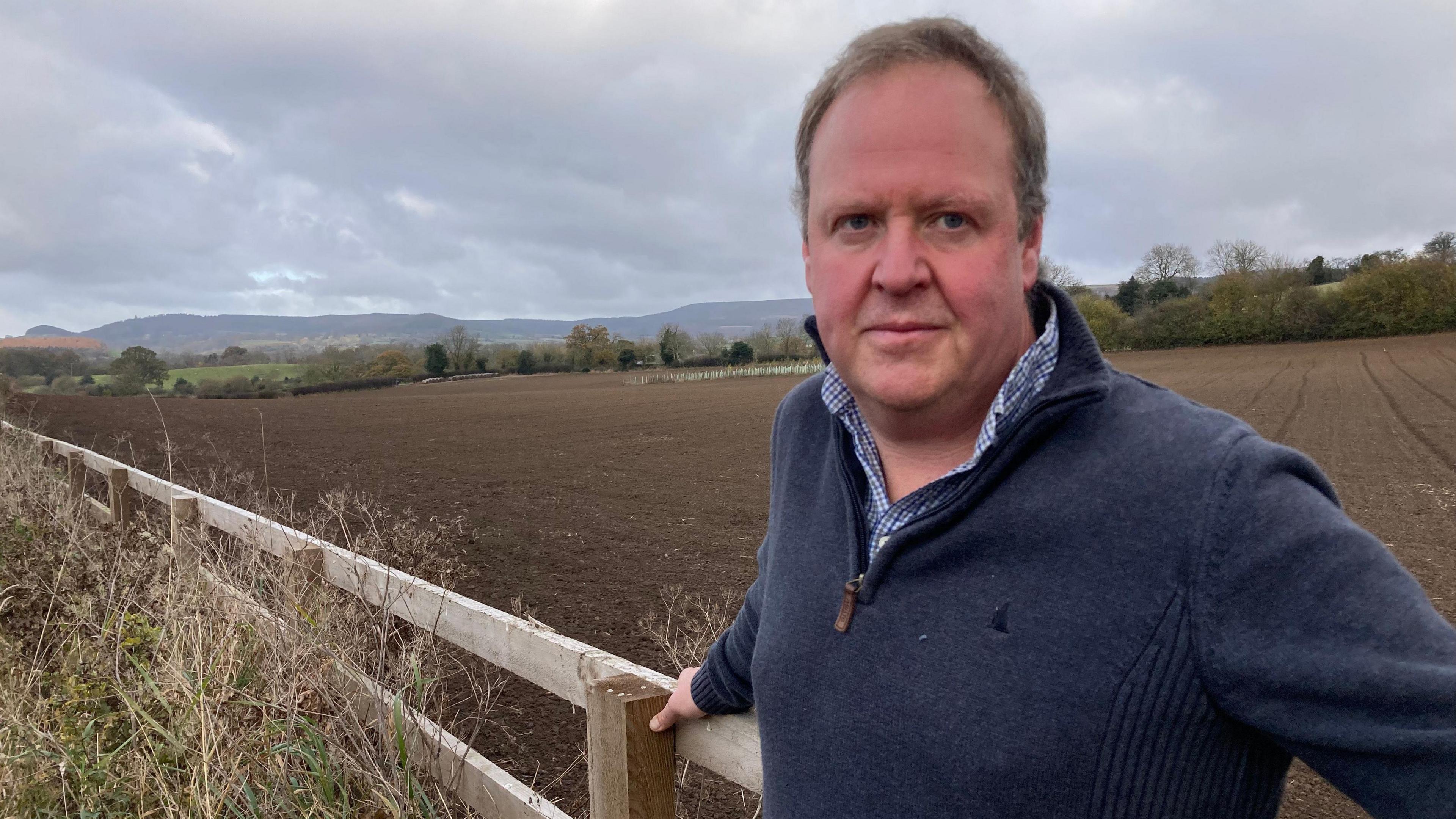 A man, wearing a navy quarter zip fleece and check shirt, leans against an outdoor wooden fence. The background is of a muddy ploughed field. 