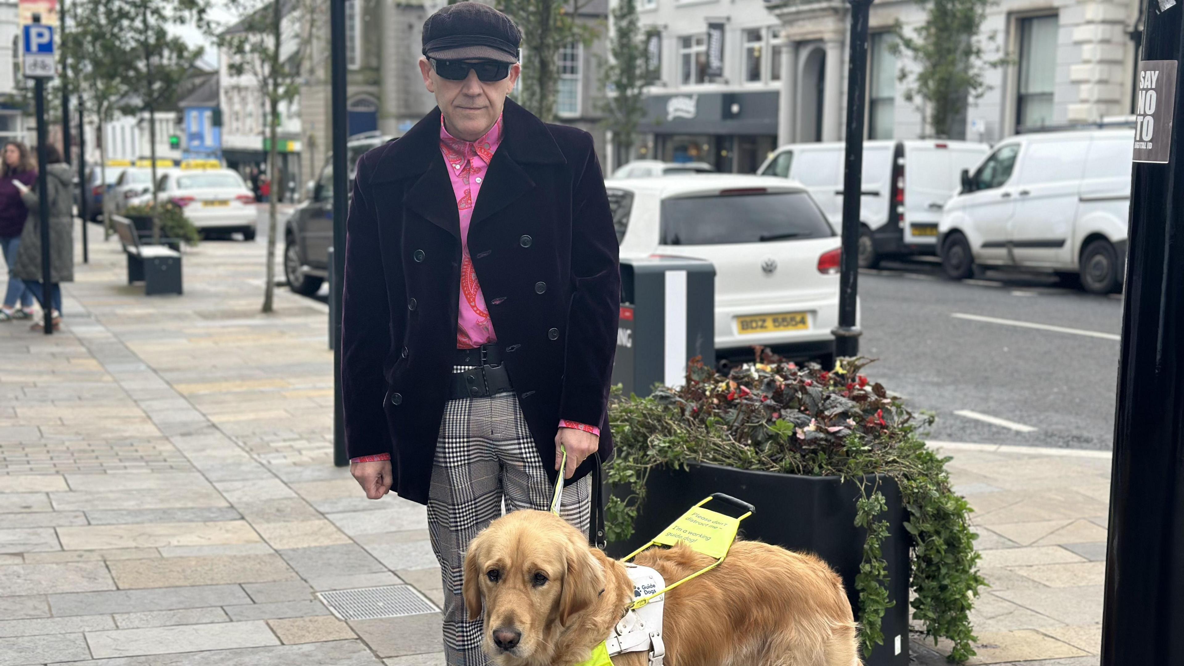 Alex is wearing dark glasses and a dark cap. He is wearing a pink shirt, purple velvet jacket and grey check trousers. Alex is standing on Main Street with his golden guide dog Angus next to him. Angus has a sign on his lead which says he is a working guide dog. They are standing next to a street planer with foliage. There are cars parked in the background, lampposts and shops in the distance.