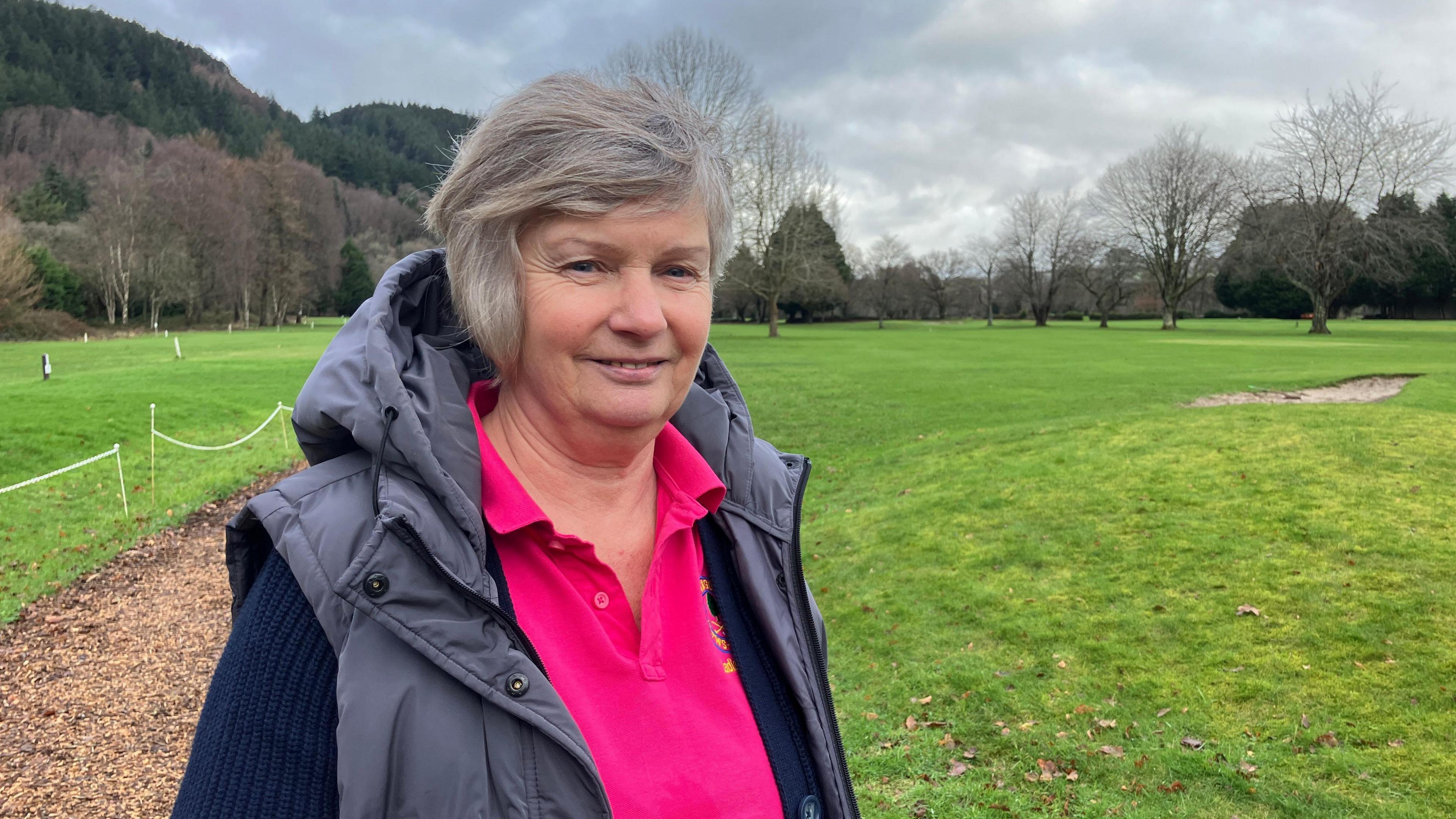 Delyth Pierce mewn crys polo pinc, cardigan glas, a gilet llwyd. Mae hi'n sefyll o flaen cwrs golff.