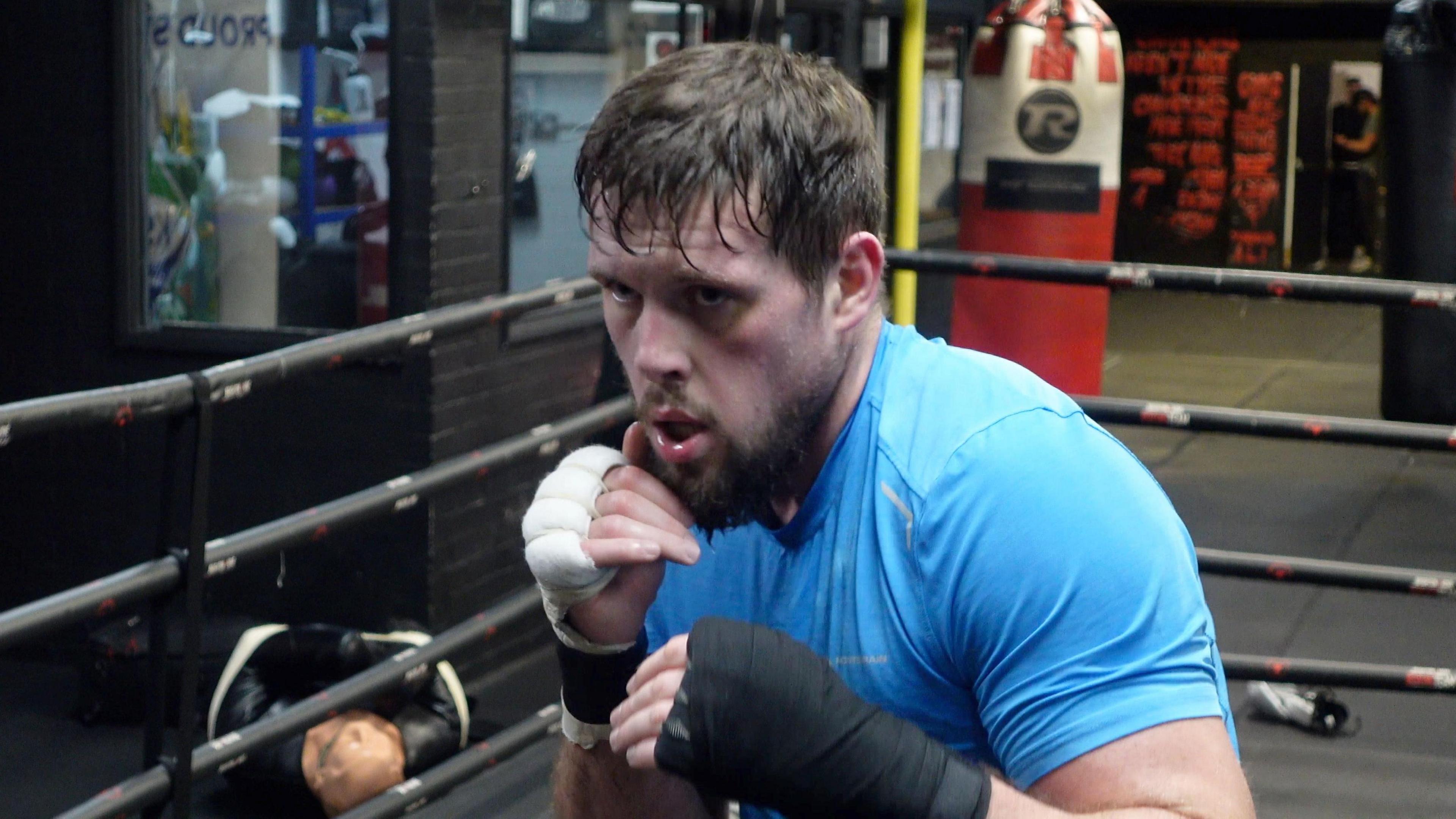 Tommy Welch, holding his fists up, while training at Brighton and Hove Amateur Boxing Gym, with ropes and a boxing bag in the background.