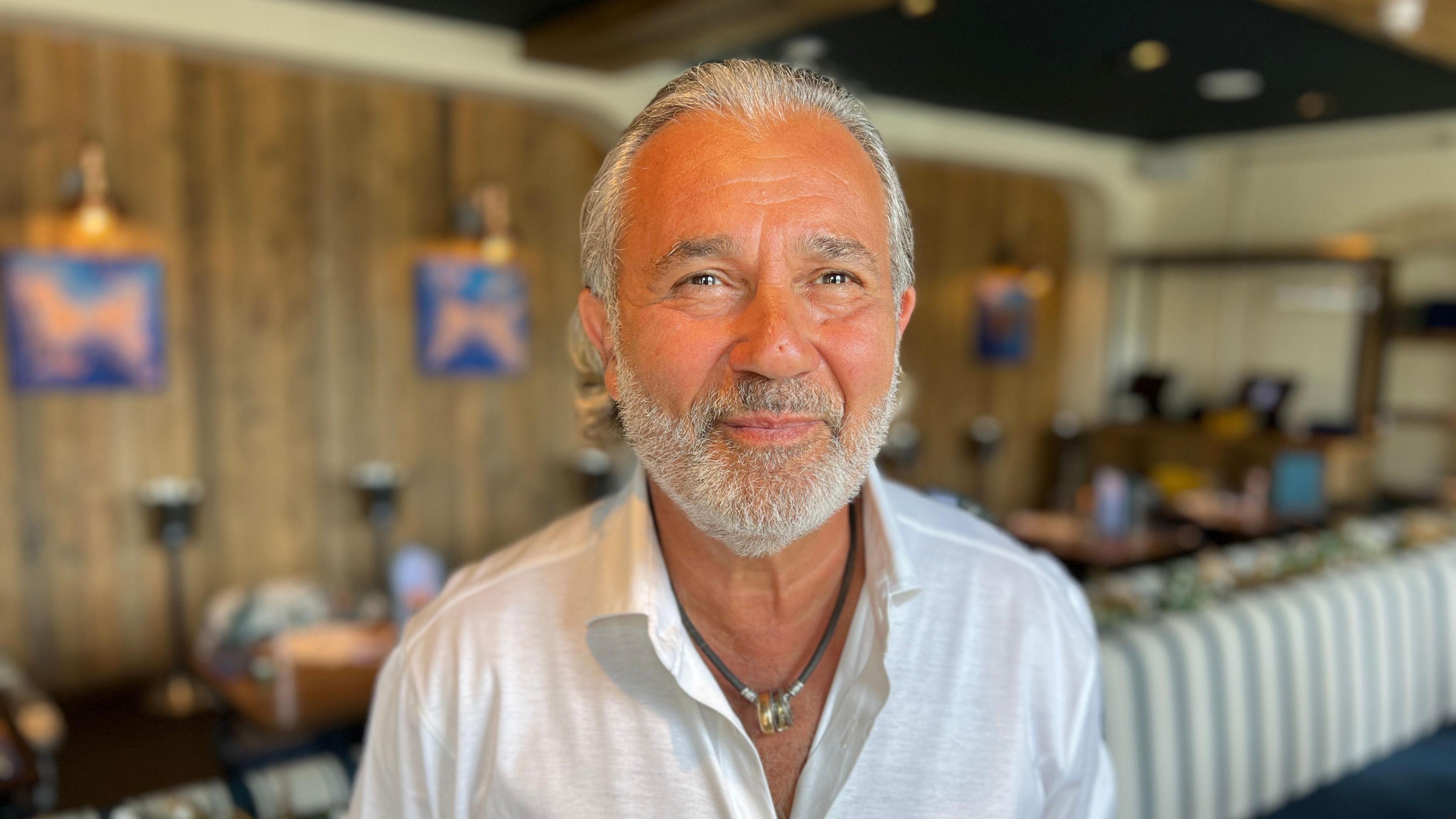A tanned middle-aged man with a white beard smiles as he looks at the camera