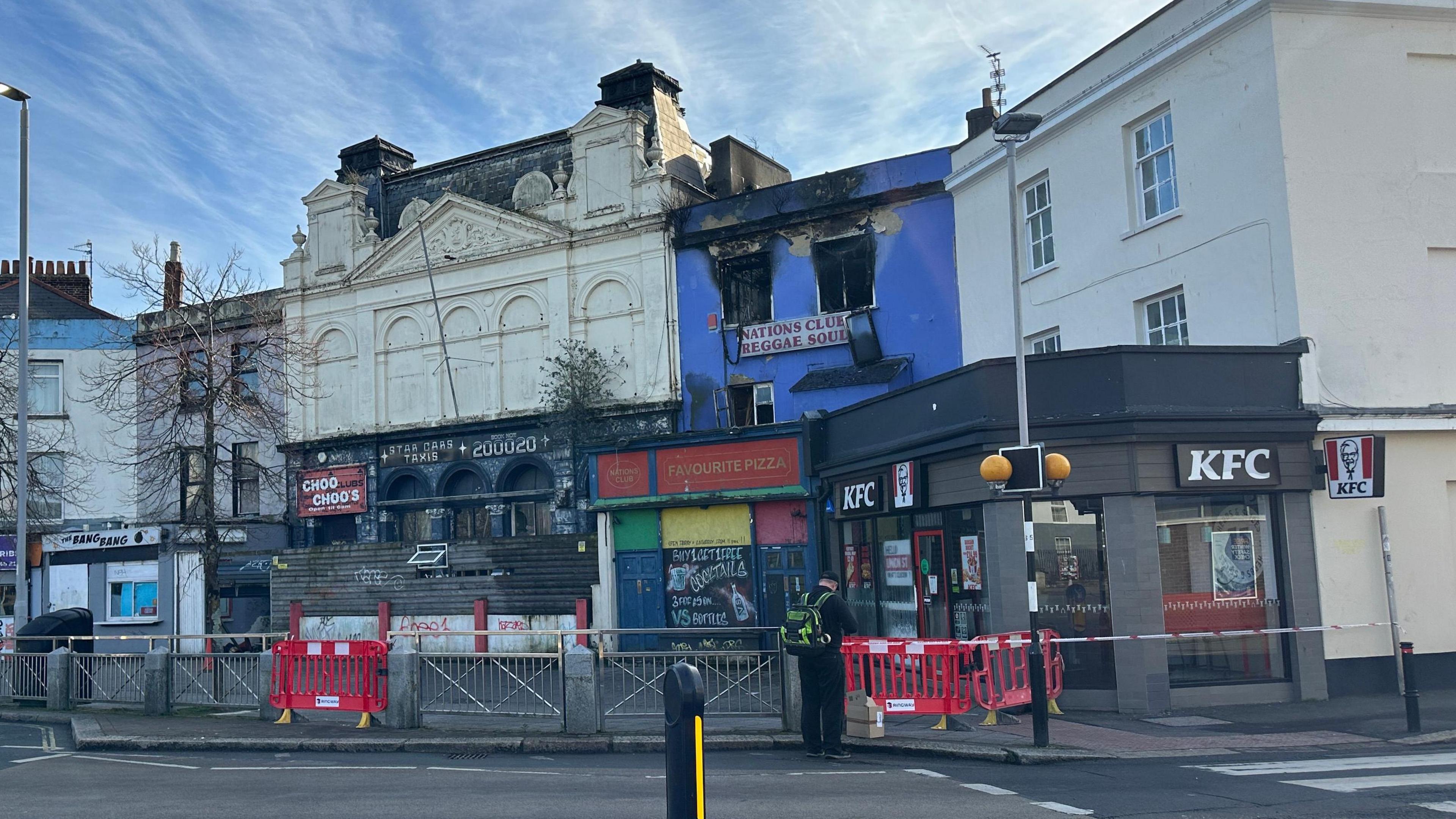 A cordon has been set up following a fire in Plymouth. A building called the Nations Club has been damaged in the blaze with its blue exterior walls charred. It is next to a KFC and another large building. Tape and red fencing has been set up around the scene. A person with a high-vis backpack is standing on the edge of the cordon.