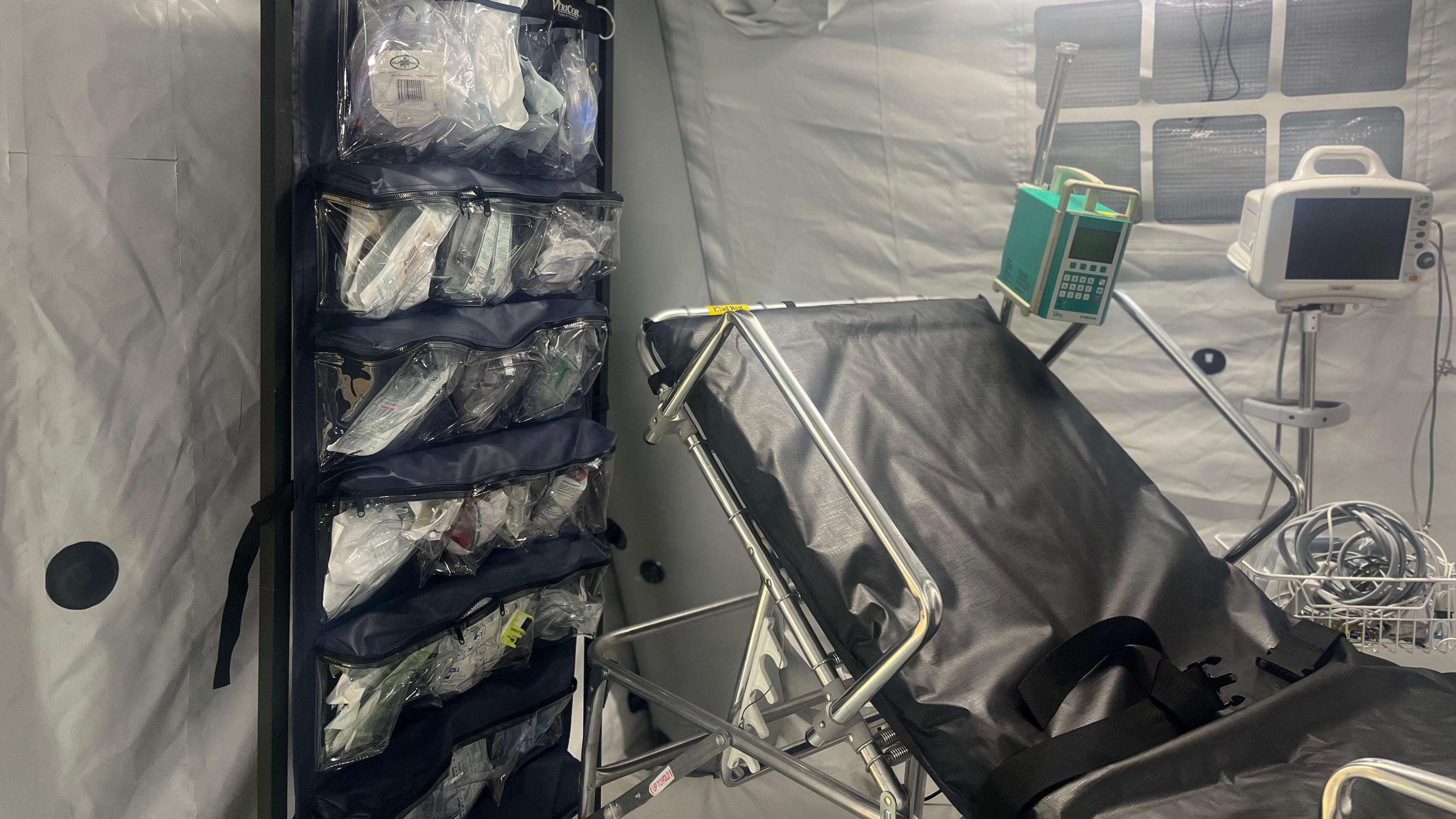 A black hospital trauma bed, basic vital sign monitors, ultrasound scanners, and ECG monitoring equipment is visible inside a white tent.
A large black and blue cabinet contains a number of plastic bags containing medical equipment.
