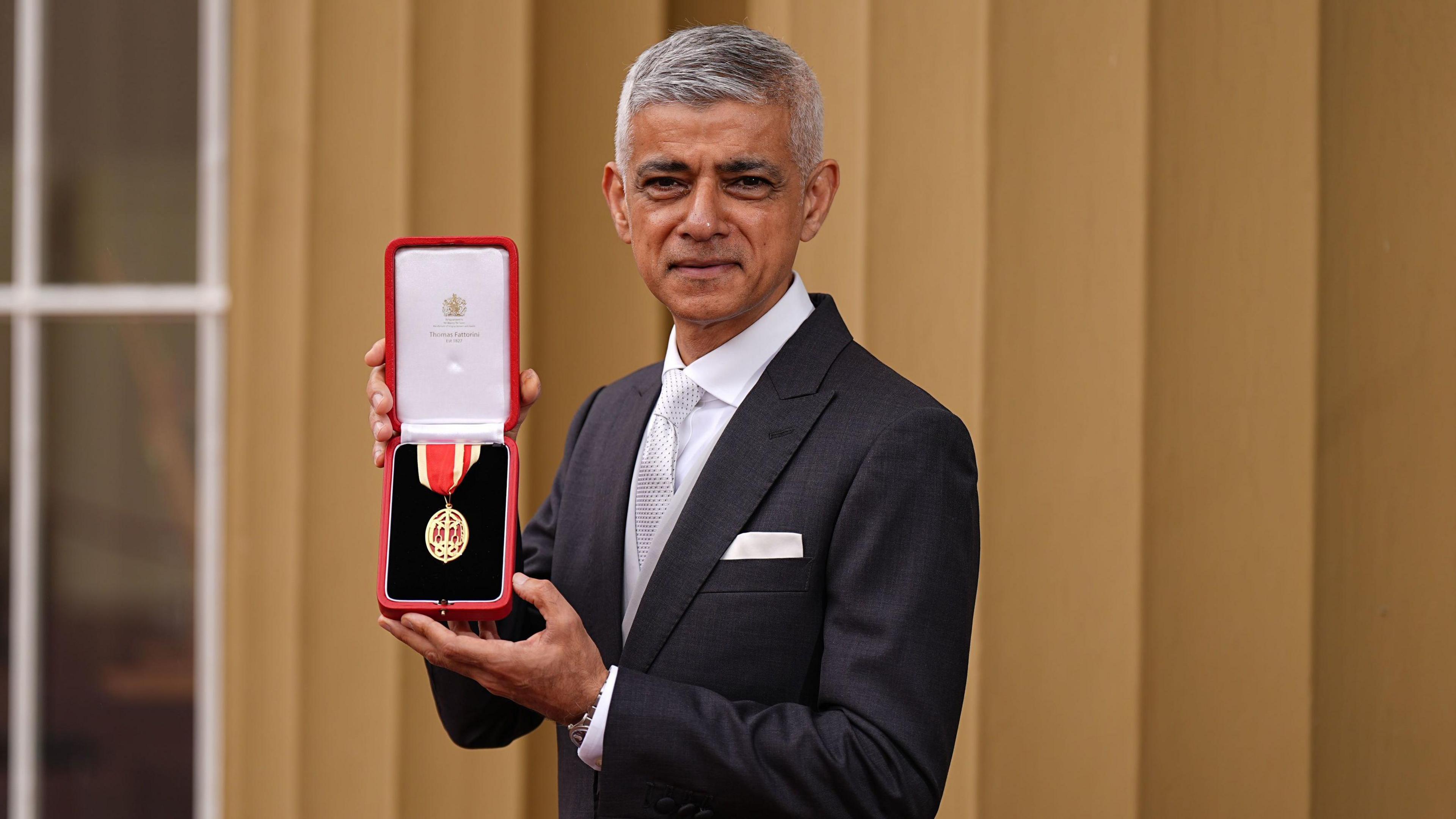 Sir Sadiq Khan in morning dress stands outside Buckingham Palace holding his medal and smiling for the cameras.