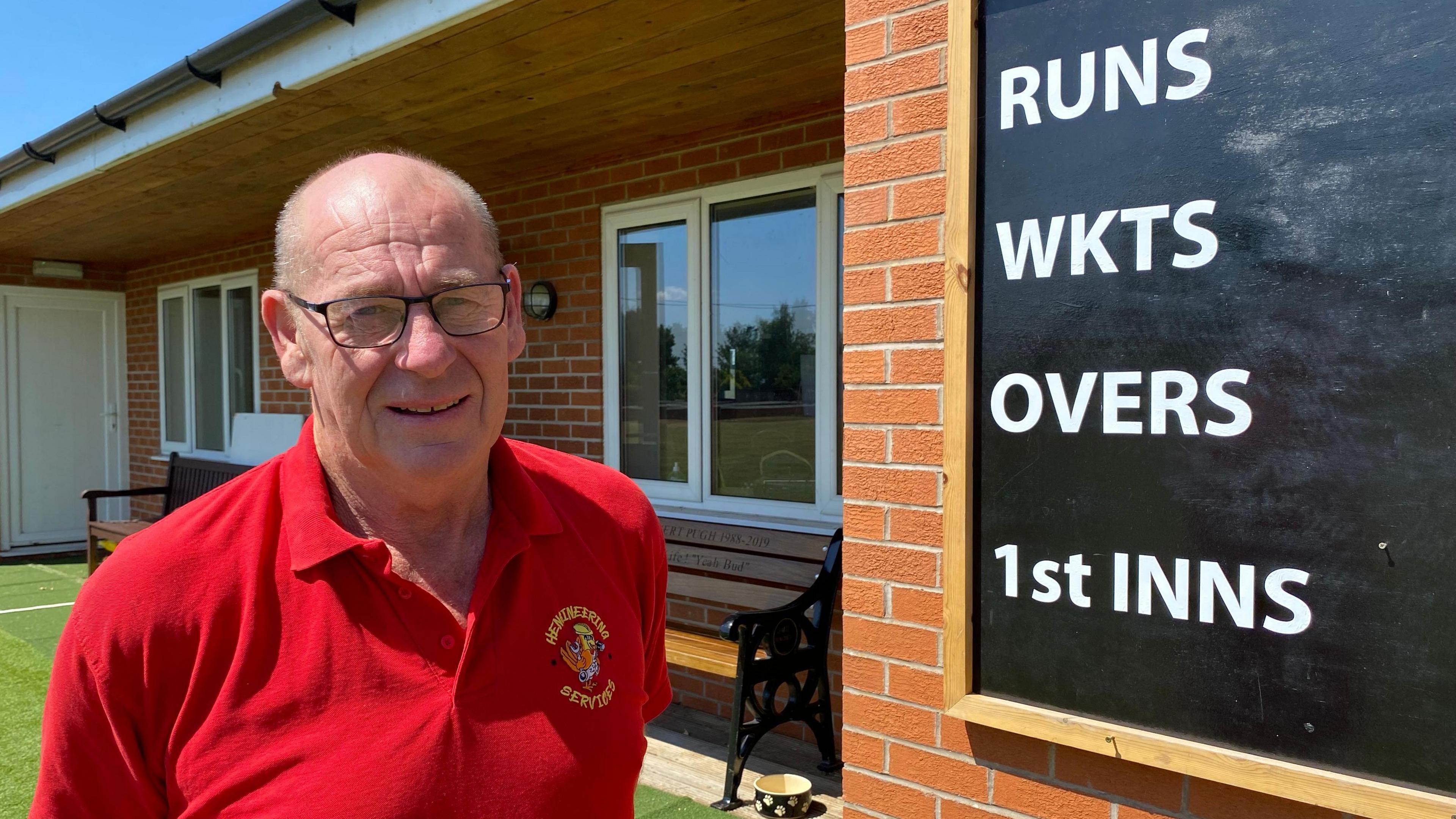 A man with very short grey hair and black classes standing outside a cricket club. There is a black board with white writing that reads "RUNS, WICKETS, OVERS and 1st innings".