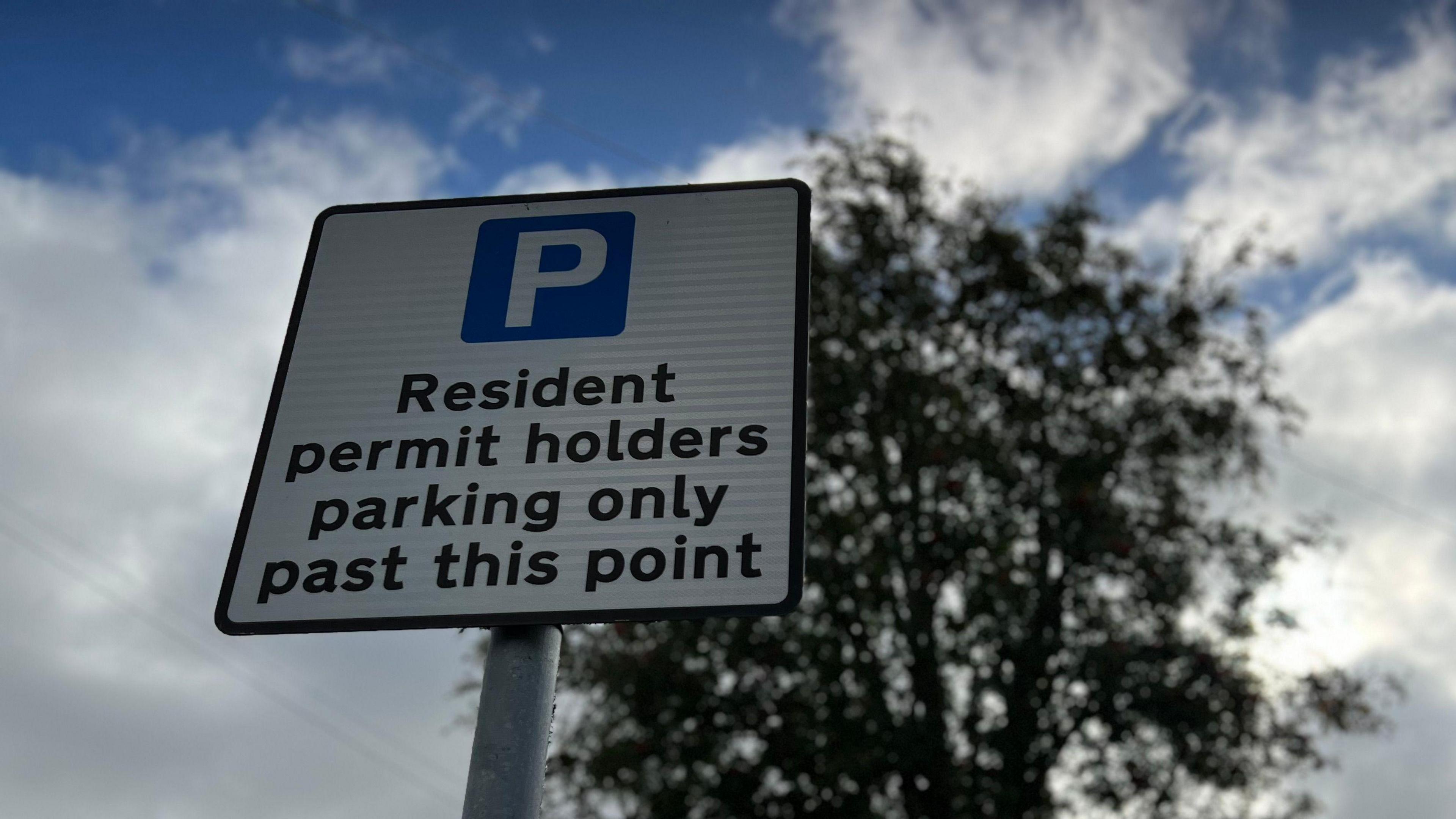 A close up photo of a resident permit parking sign. It is taken from a low angle looking up at the sign with a tree in the background on a cloudy day.