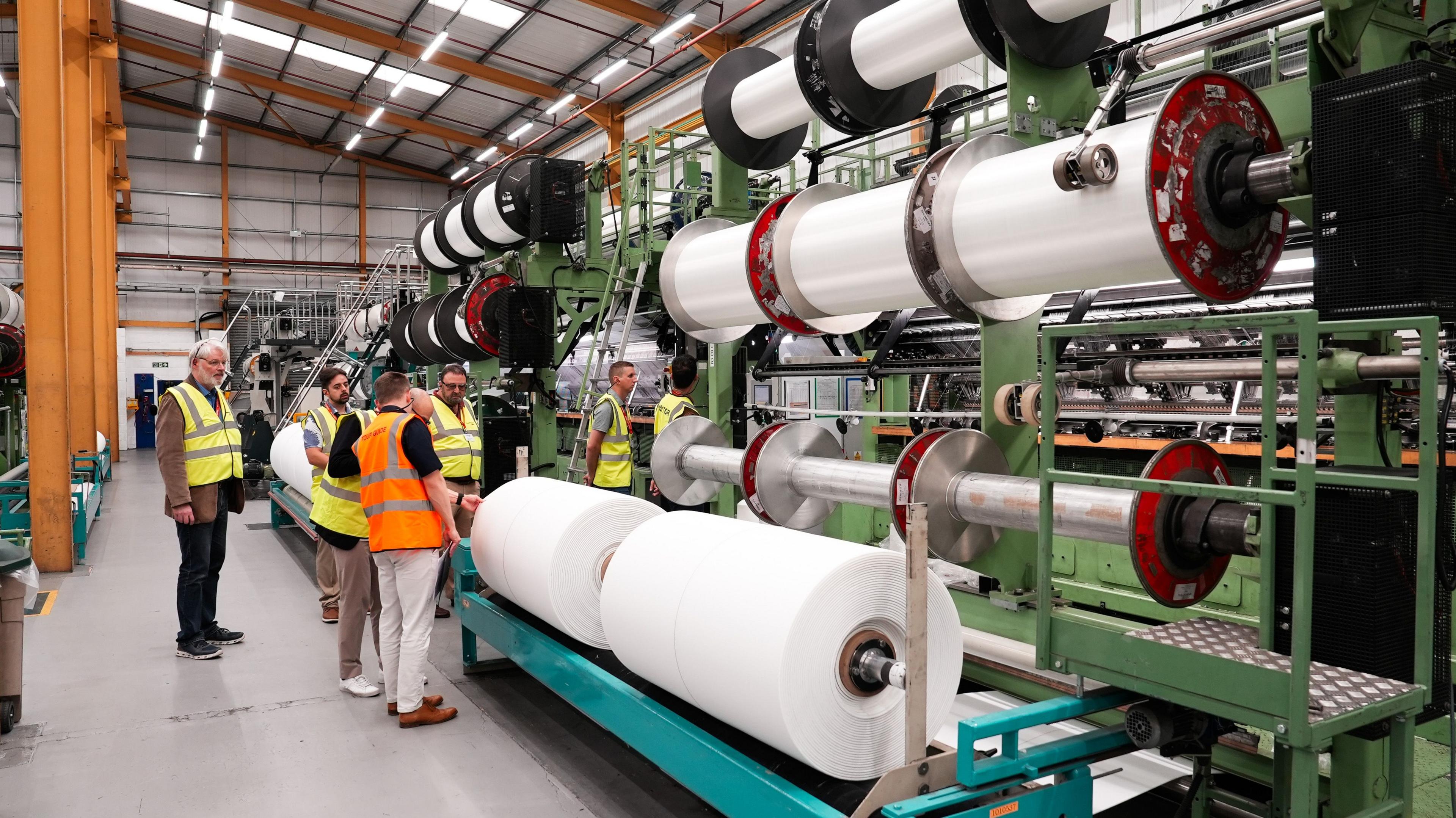 Huge reels of yarn are seen stacked on spools to the right while on the left delegates examine them. 