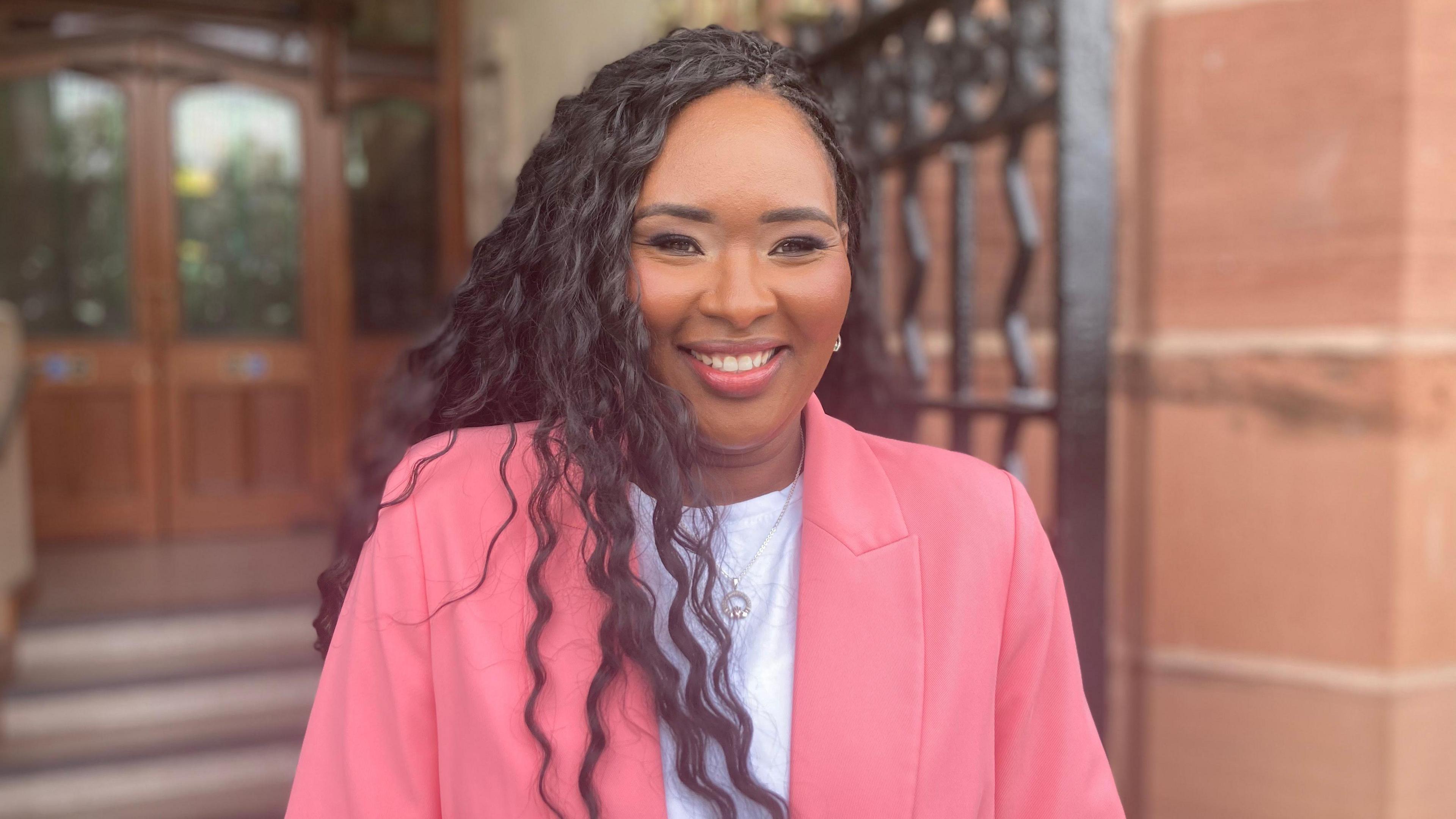 Lilian Seenoi-Barr is smiling into the camera. She is standing in fron of a stone entranceway and a black metal gate. She is wearing a white top and a pink blazer 