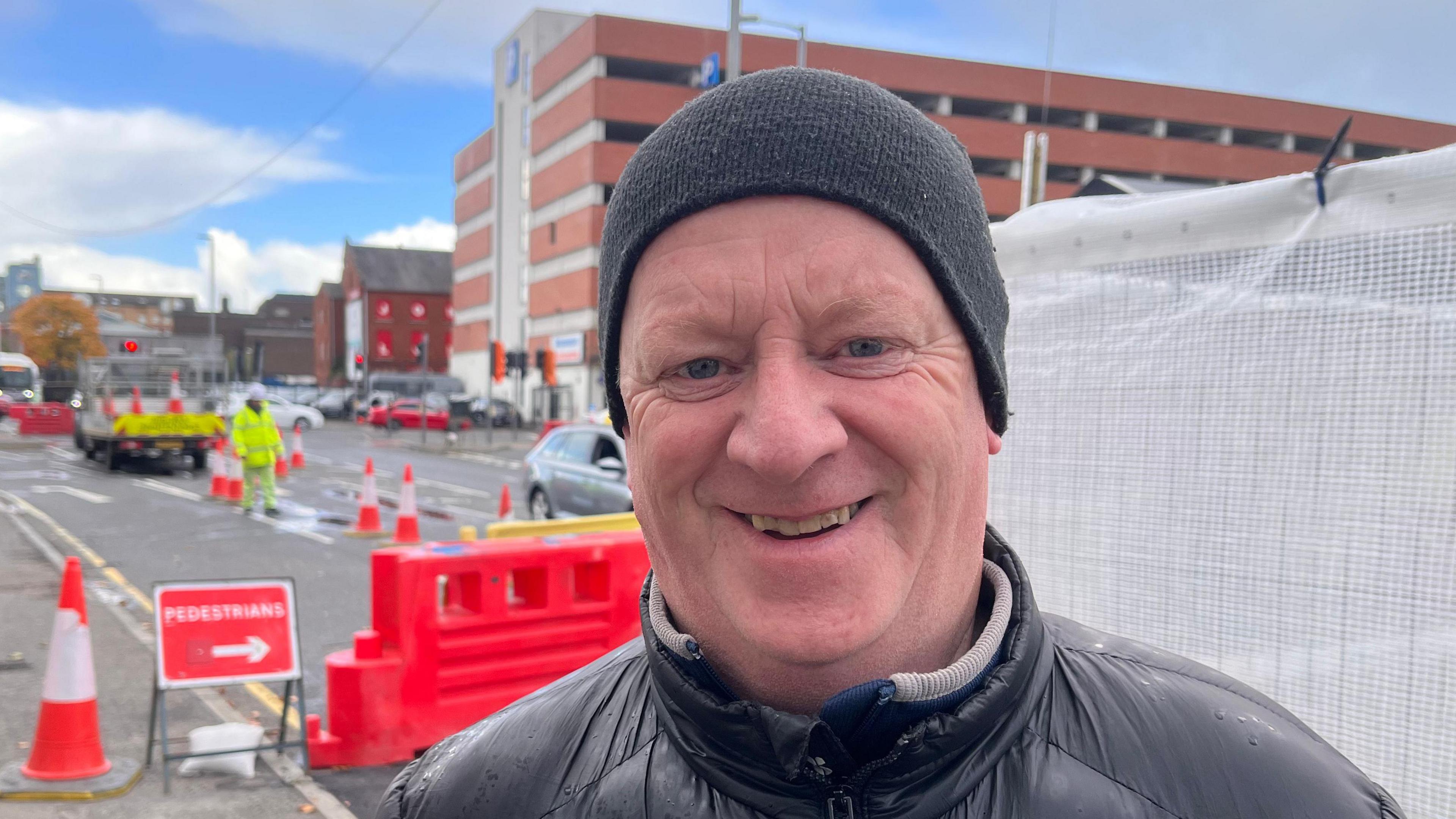 Christopher Douglas. He is wearing a black cap and black puffer jacket. Roadworks can be seen in the background.